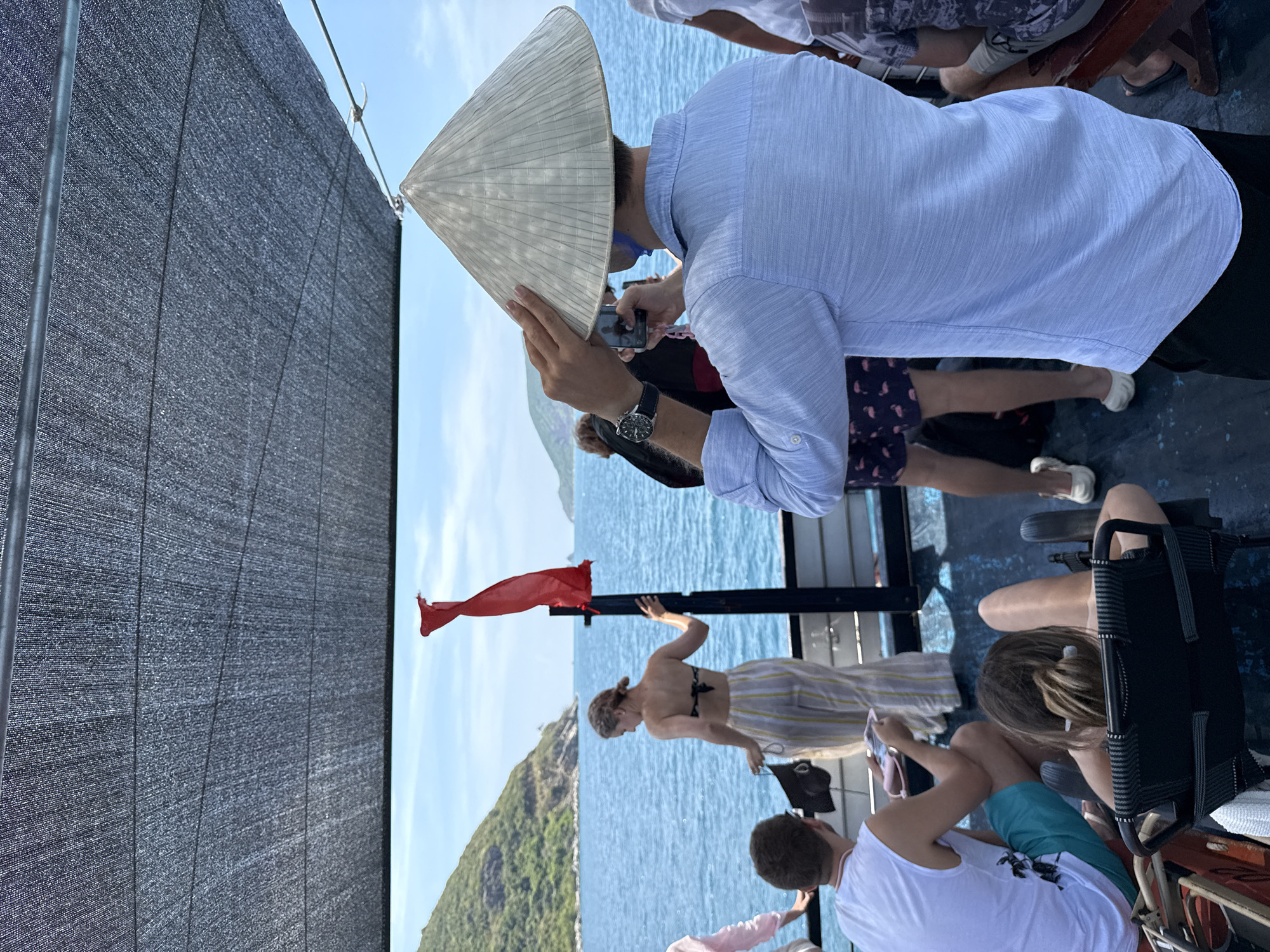 The image depicts a group of tourists on a boat in what appears to be Nha Trang Ward, Vietnam, based on the presence of a traditional Vietnamese conical hat worn by a man taking a photograph. Another person in a bikini and striped pants poses by a red flag attached to a black metal pole, possibly on a tourist boat or ferry. The background shows a hilly coastline, the sea, and a clear sky. Other people are visible, seated or standing on the boat. A black sunshade is partially visible on the left side of the image.