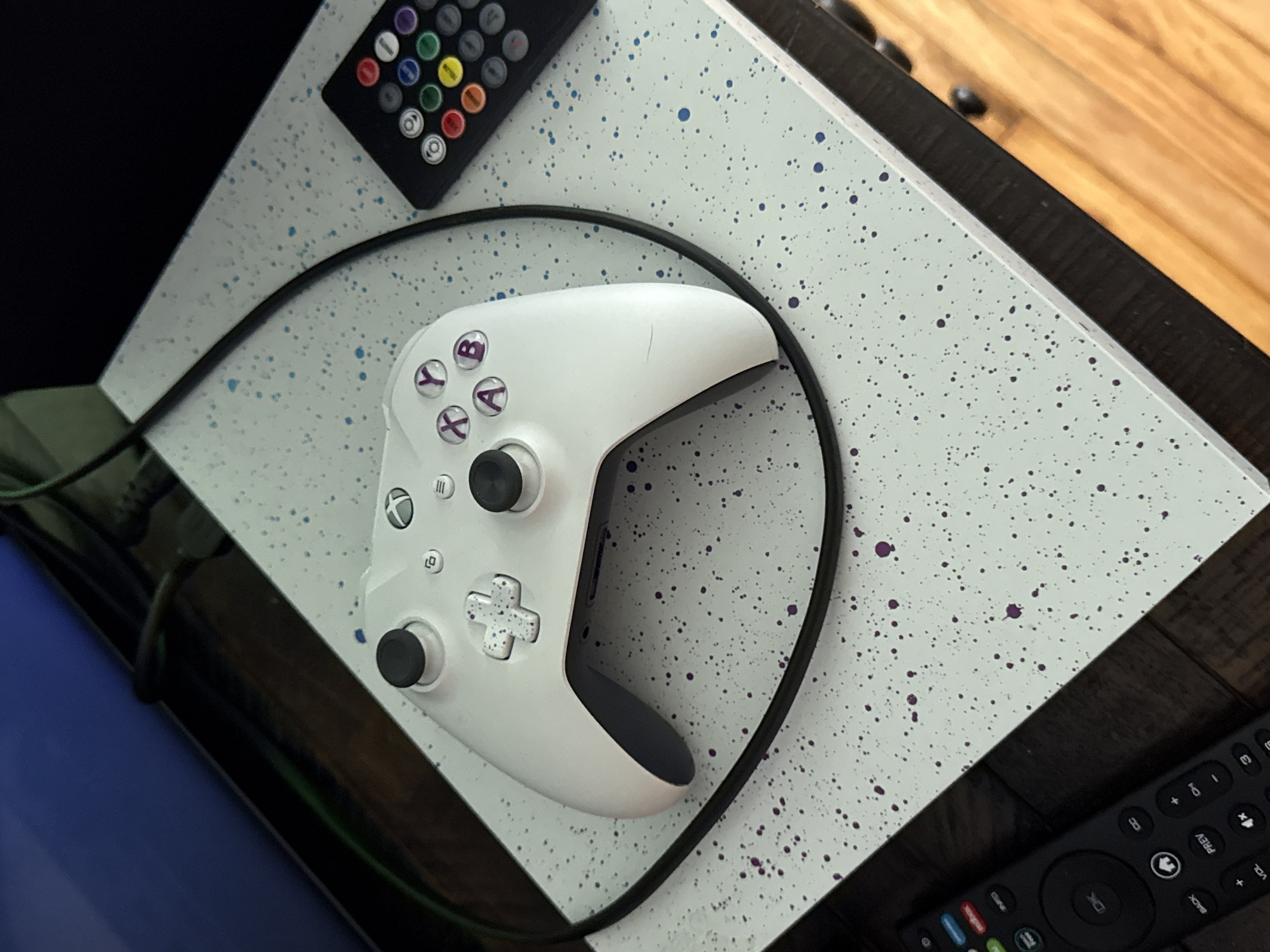 In an indoor setting, an Xbox controller and a remote control are placed on a white, speckled surface. The Xbox controller, which is white with purple buttons, sits in the center of the frame. To the right of the controller is a black remote control with numerous colored buttons. A black cord snakes around the controller and extends out of frame to the right. The surface the objects are on is light blue with dark blue and purple speckles. The background is dark and out of focus, with hints of a wooden floor visible in the lower right corner. The scene is forensic and matter-of-fact, with no people or interactions depicted. The location is not specified.