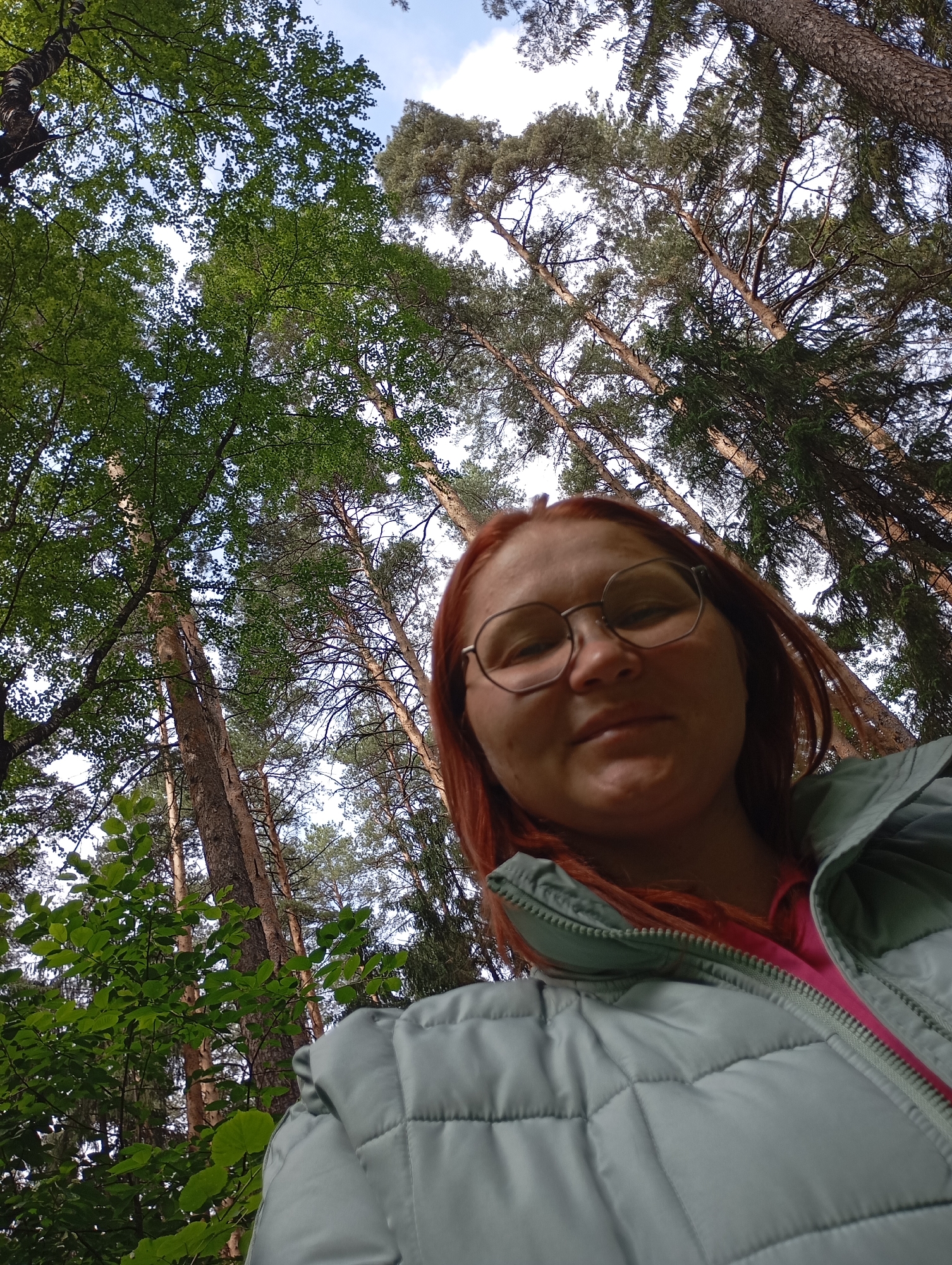 A low-angle selfie captures a woman in a forest setting in Chaikovsky, Russia. The woman, with red or auburn hair, is positioned in the lower half of the frame, looking slightly downwards towards the camera. She wears round, wire-frame glasses and a light blue or mint green quilted puffer jacket, featuring a bright pink interior lining visible at the partially unzipped collar. Her expression is neutral, with a subtle hint of a smile.
The background is dominated by a dense forest canopy, composed of tall trees stretching upwards. Both deciduous trees with lush green leaves and coniferous trees with needle-like foliage and textured bark are visible. Glimpses of a bright, partly cloudy sky with patches of blue are seen through the gaps in the branches, indicating daytime, likely late morning or early afternoon. The scene conveys a sense of being outdoors amidst nature. No visible text is present.