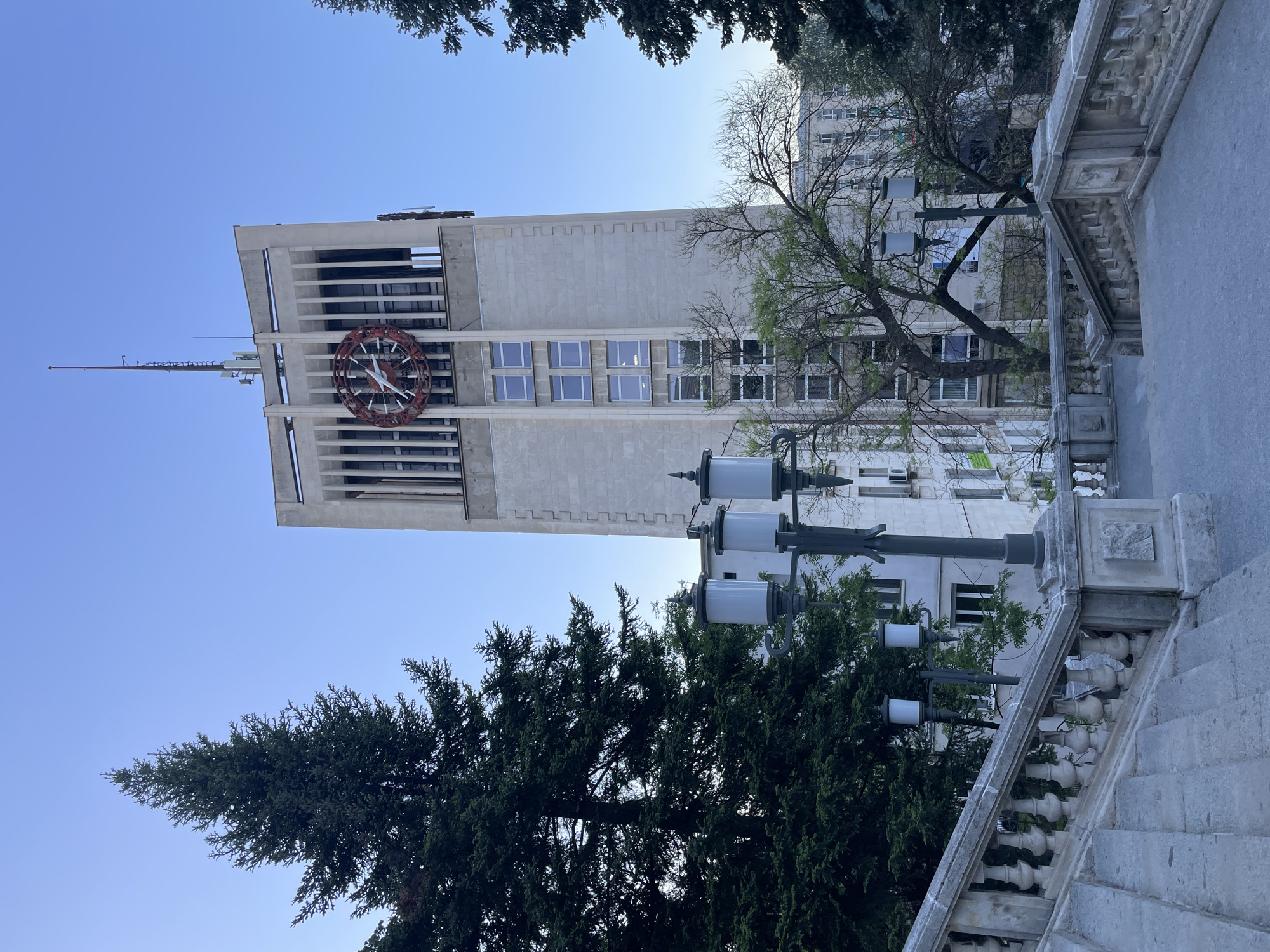 The image depicts the Gabrovo Municipality building in Bulgaria, viewed from a low angle. The structure is characterized by its clock tower, featuring a large clock face. The tower is constructed of light-colored stone or concrete and exhibits a modernist architectural style. The sky is a clear blue. Foreground elements include trees with dense green foliage and a section of ornate stone steps and a railing. A multi-bulb lamppost stands near the steps. A building with numerous windows is partially visible to the right of the tower.