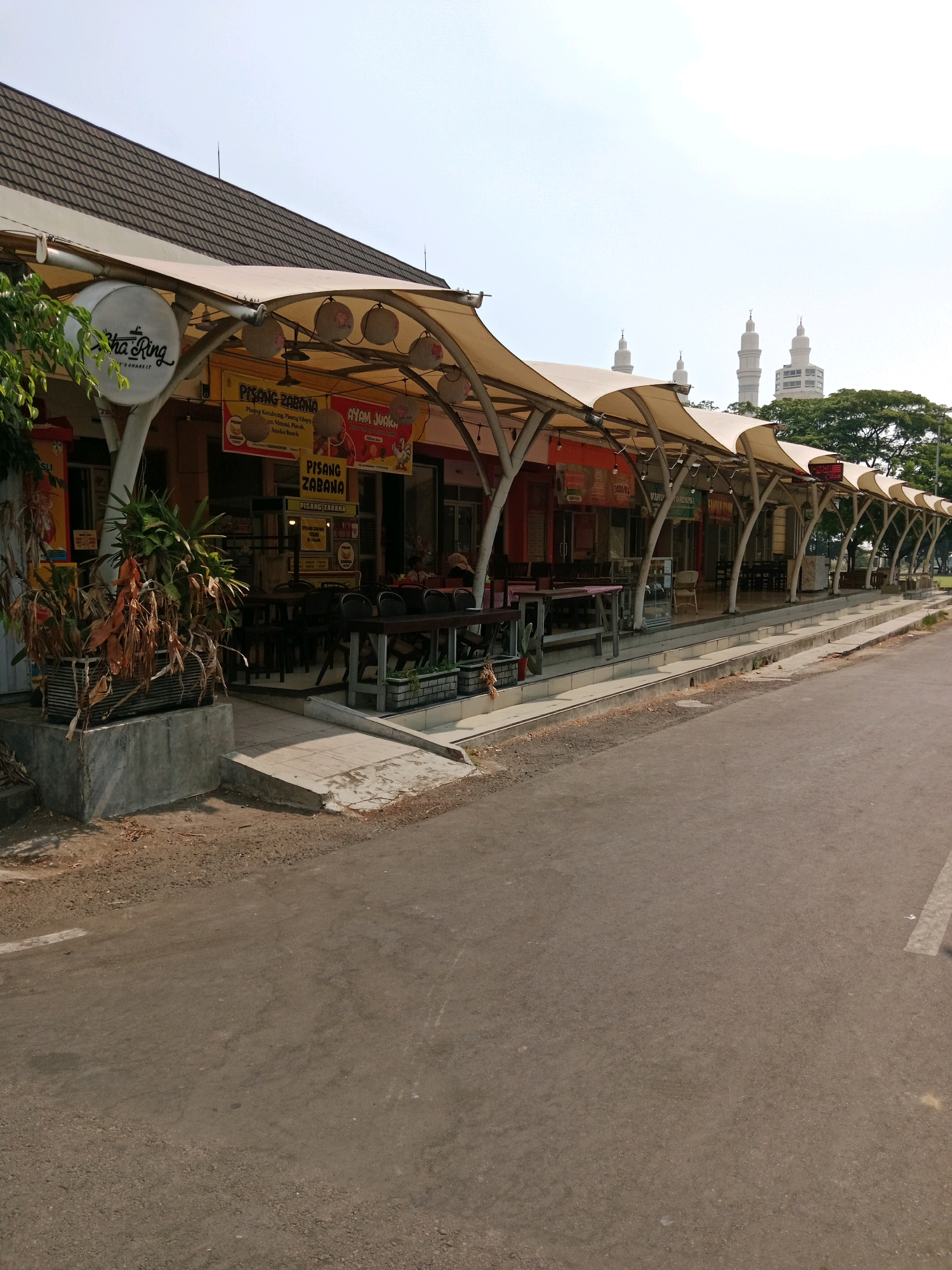 This is a medium-shot, eye-level, outdoor, daytime photograph of a row of food stalls with a canopy in the city of Karanganyar, Indonesia. The stalls are built along a paved road with a sidewalk. The buildings behind the stalls are mostly single-story with a dark-tiled roof. A white canopy extends over the food stalls, supported by metal poles. Signs on the stalls advertise various food items. In the background, several white minarets of a mosque are visible, along with trees and a tall building. The road is asphalt and appears to be in good condition. There are no people visible in the immediate vicinity of the stalls.