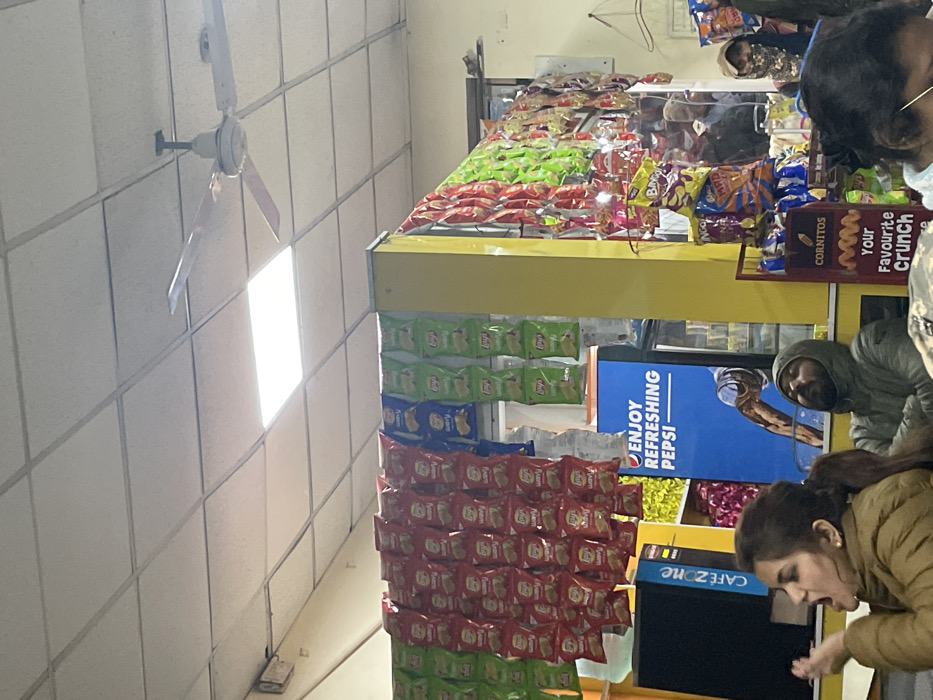 The image depicts the interior of a snack shop or canteen, likely in Haldwani, India, bustling with activity. The setting features a white tiled ceiling with a bright rectangular light fixture and a partially visible white three-bladed ceiling fan.
The shop is well-stocked with a variety of packaged snacks displayed on yellow and black shelving units. Bags of chips, including Lay's (in green, blue, and red packaging), Bingo!, and Cornitos, are prominently arranged on shelves and hanging racks.
Several people are present in the shop. In the foreground on the right, a woman with dark hair, wearing a light brown jacket, is bent over, looking intently at something at a lower level, possibly selecting an item or using a machine. Behind her, a man with a beard and a grey hooded jacket stands, looking towards the shop's front. Other individuals are partially visible in the background, some behind the counter, engaged in various transactions or observations.
Notable visible text includes a blue poster with "ENJOY REFRESHING PEPSI," a red sign for "CORNITOS Your Favourite Crunch," and "CAFE ZONE" on a black machine, likely a beverage dispenser or coffee maker. The scene suggests a busy retail environment, consistent with a daily operation. The time of day and weather are not discernible from the indoor lighting.