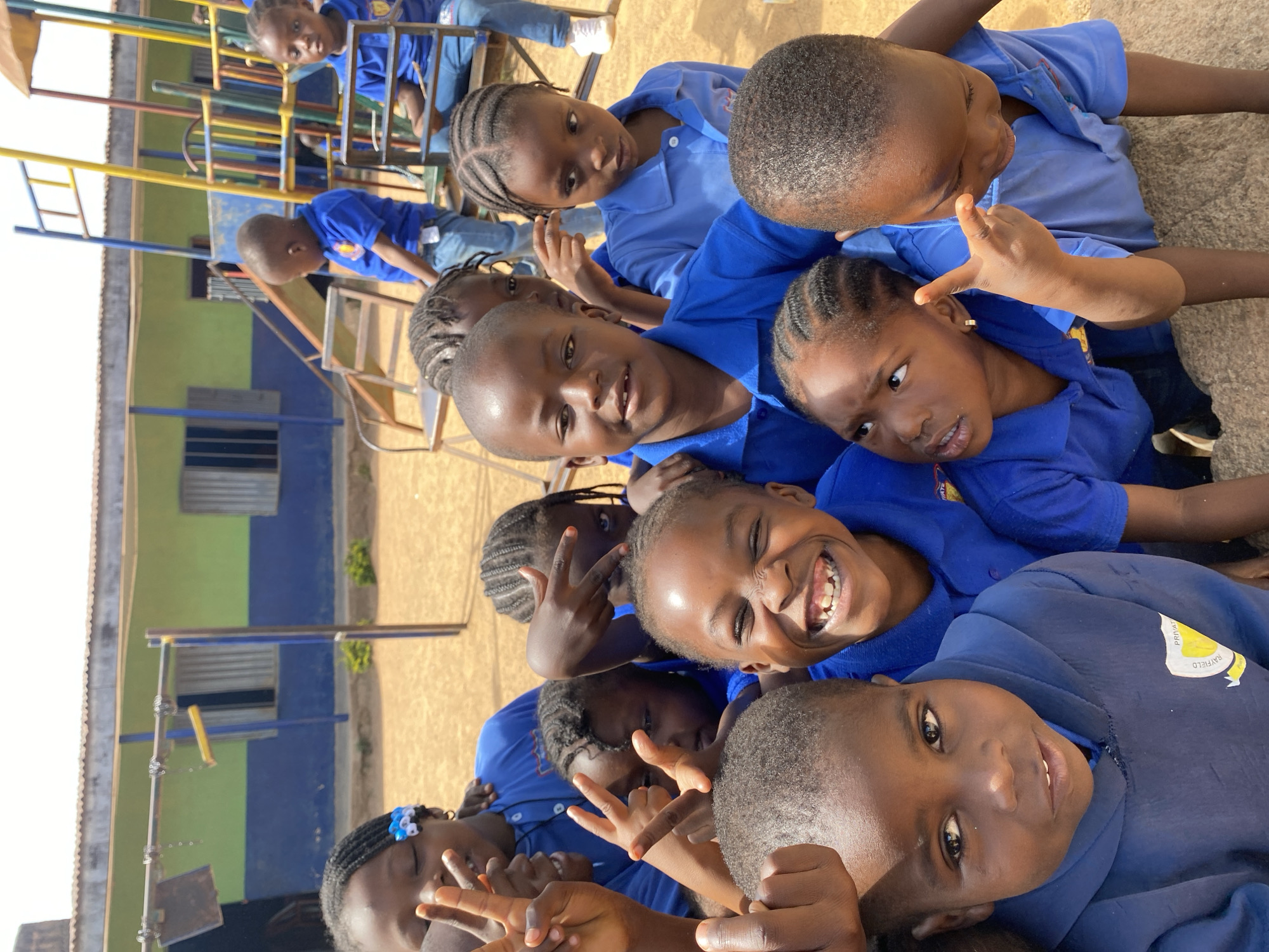A group of children, mostly girls, wearing blue uniforms, are huddled together on an outdoor surface, possibly concrete, in Gura Top, Nigeria. Some are making peace signs with their hands and smiling at the camera. A playground structure with blue, yellow, and red elements and a blue and green building are visible in the background. Some other children in blue uniforms are visible on the playground structure.