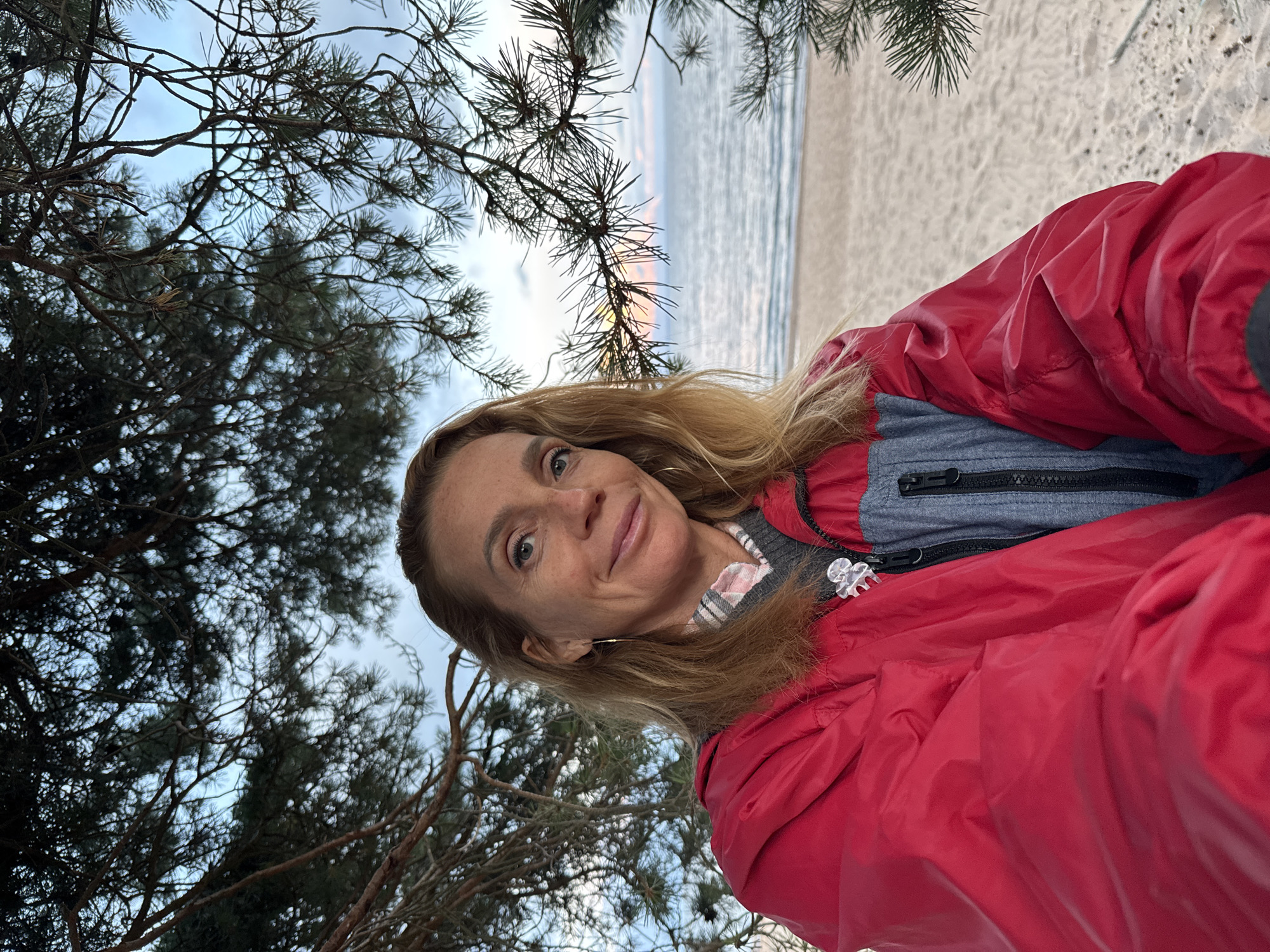 A woman with blonde hair and blue eyes is pictured outdoors, likely near the sea, with pine trees framing the shot. She is wearing a red jacket over a gray and white plaid shirt and a dark grey sweater. A silver-colored brooch is attached to the left side of her red jacket. The background shows a sandy beach, the sea, and a cloudy sky. The image appears to have been taken at dusk, given the lighting conditions. The location is identified as Shepelevo, Russia, suggesting a coastal setting in that region.