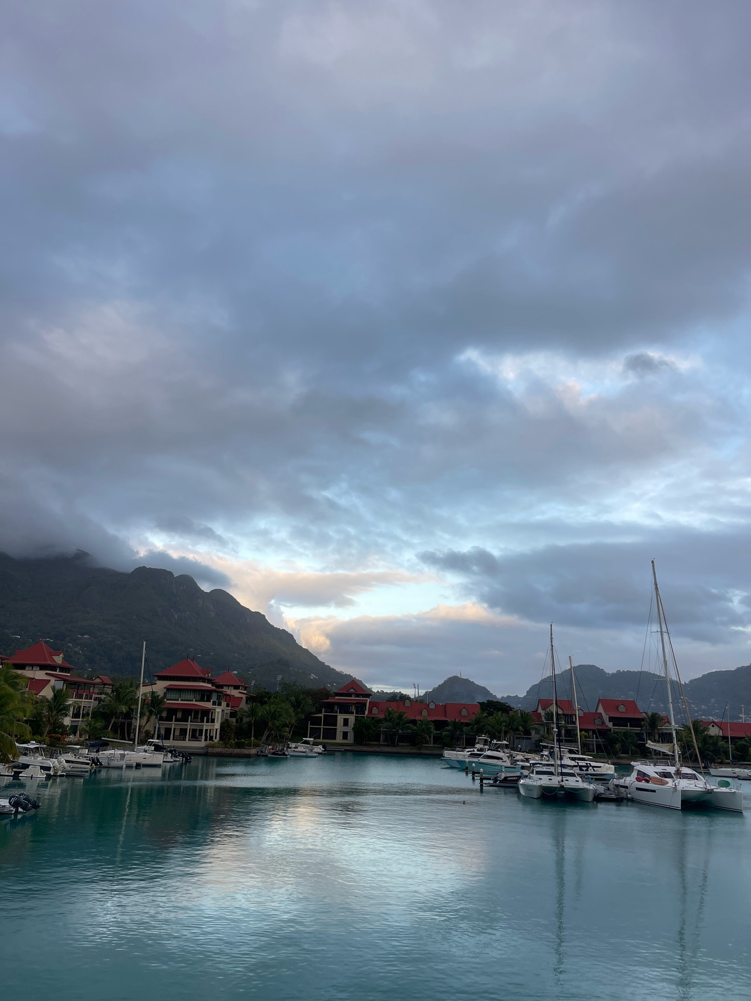 The image shows a harbor in Victoria, Seychelles, with a calm, turquoise body of water in the foreground. Numerous boats, including sailboats and smaller motorboats, are docked along a waterfront lined with buildings. These buildings have distinctive red-tiled roofs and are surrounded by palm trees. In the background, lush green mountains rise, partially obscured by a dramatic, cloudy sky. The overall atmosphere is serene, with the reflections of the boats and buildings visible on the water's surface.