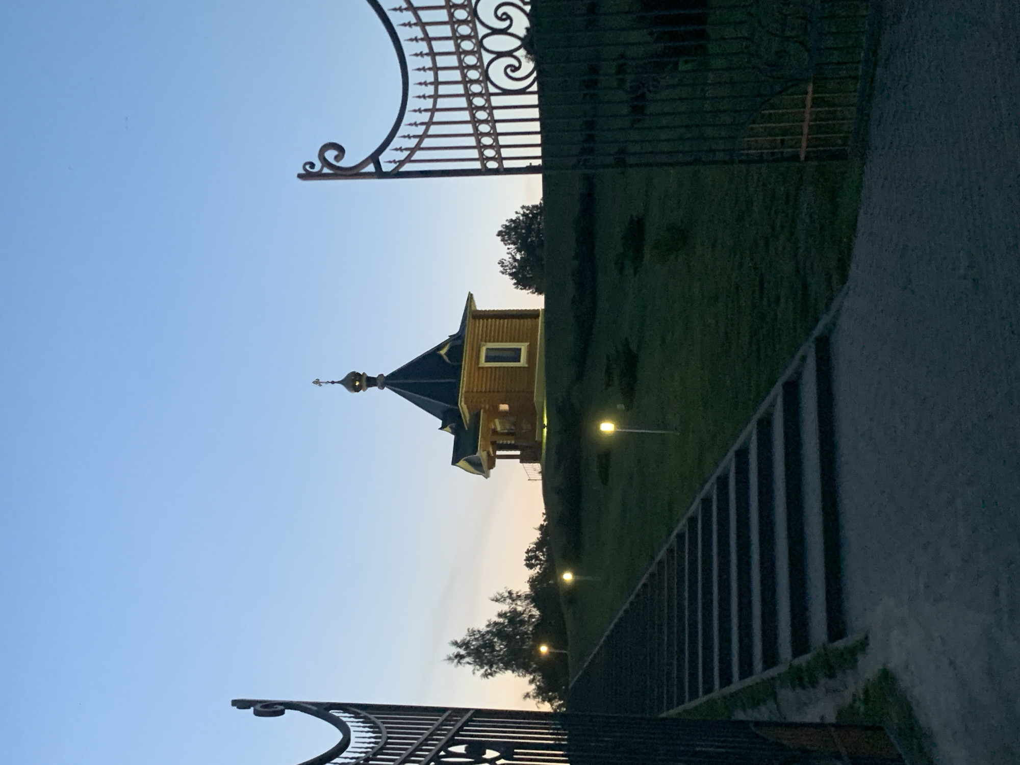 The image captures a wooden chapel with a golden dome, situated atop a grassy hill under a twilight sky. The chapel features a dark, peaked roof with a small window. A steep staircase, illuminated by several pathway lights, leads up the hill towards the chapel. The scene is framed by ornate, dark metal gates on the left and right sides of the foreground. The ground in front of the stairs is a path of gravel and dirt, with grass on either side. Trees are visible on the hill behind the chapel, and the sky is a gradient of pale blue and orange, suggesting either dawn or dusk. The image appears to be taken in Kapyĺ, Belarus, given the context. There are no people or animals visible in the frame.