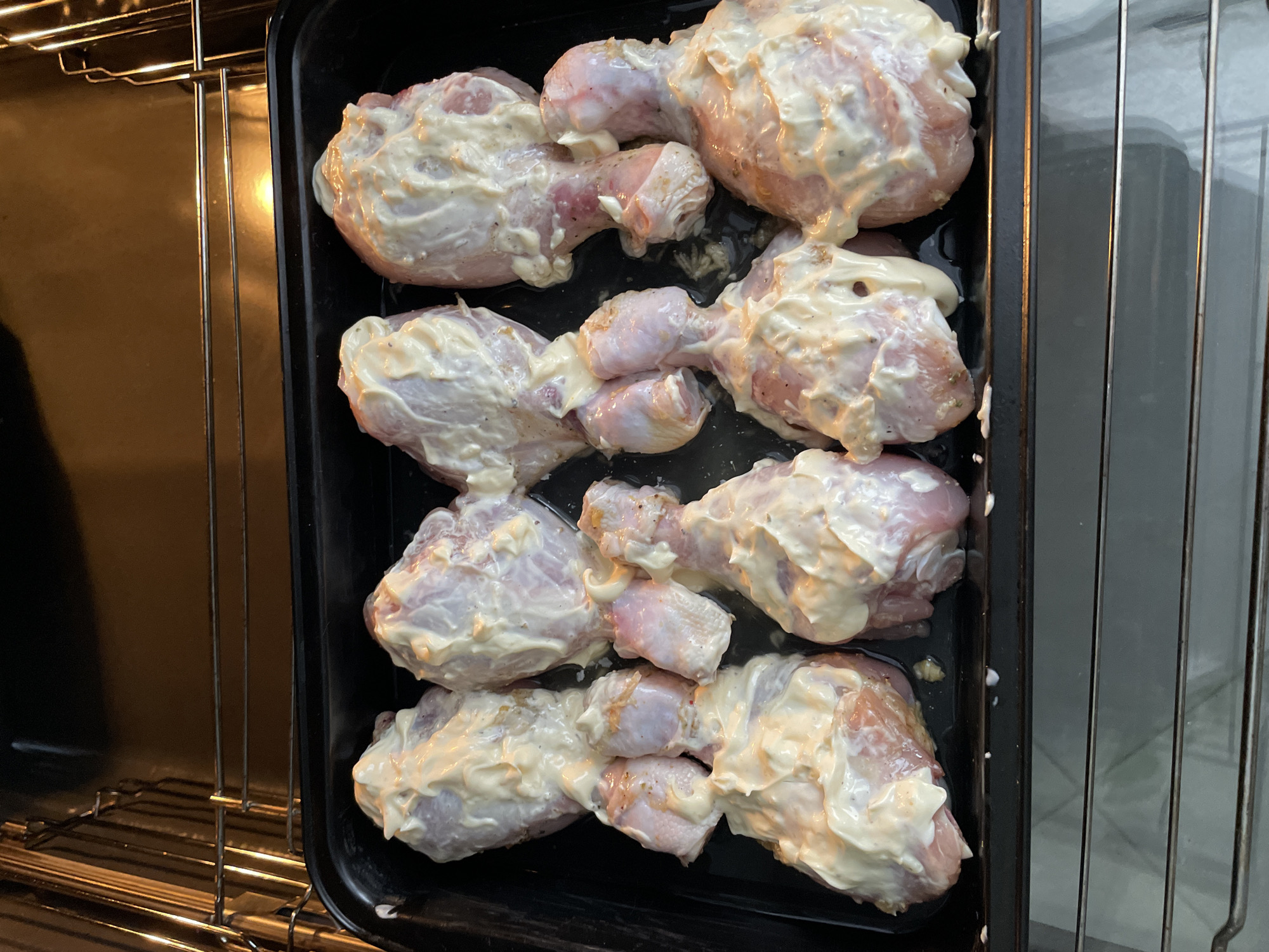 The image shows a black baking tray containing eight chicken drumsticks coated in a creamy white sauce, placed inside an oven. The oven racks are visible on the left. The location Лесной Пейзаж Кп (шарапово, Russia is extraneous and irrelevant to the image content.