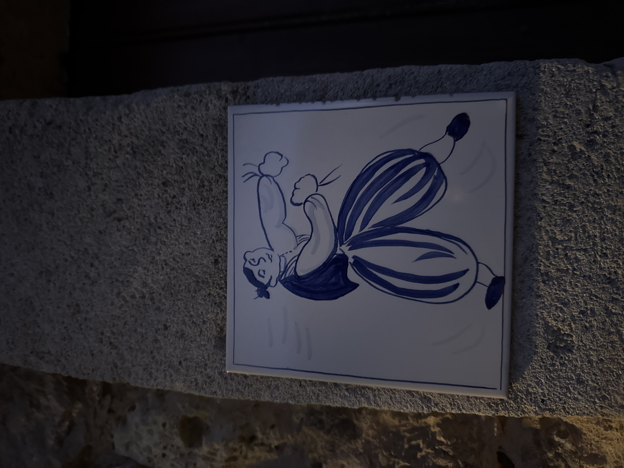 A ceramic tile depicting a person in blue and white is affixed to a rough, stone wall. The tile is square, and the artwork shows a figure with striped pants and a hat, oriented sideways. The wall is made of rough stone and appears to be exterior. The photo was taken in Valldemossa, Spain.