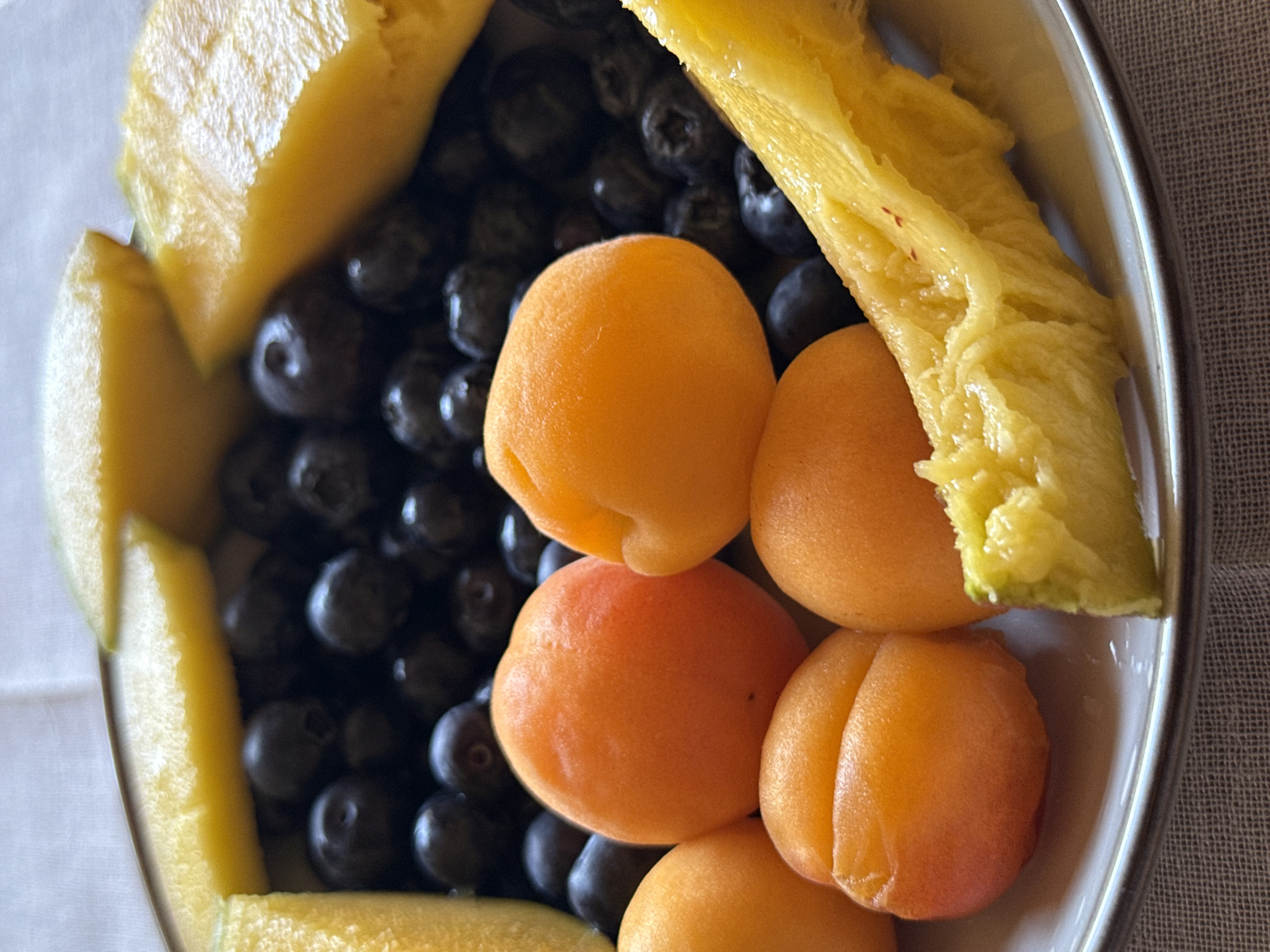 A top-down view of a shallow, oblong dish containing fruit. Yellow melon pieces are placed along the sides of the dish, with a pile of dark blueberries filling the center. Several bright orange apricots are arranged on top of the blueberries. The dish is likely sitting on a table or surface with a visible texture. The location is unknown.