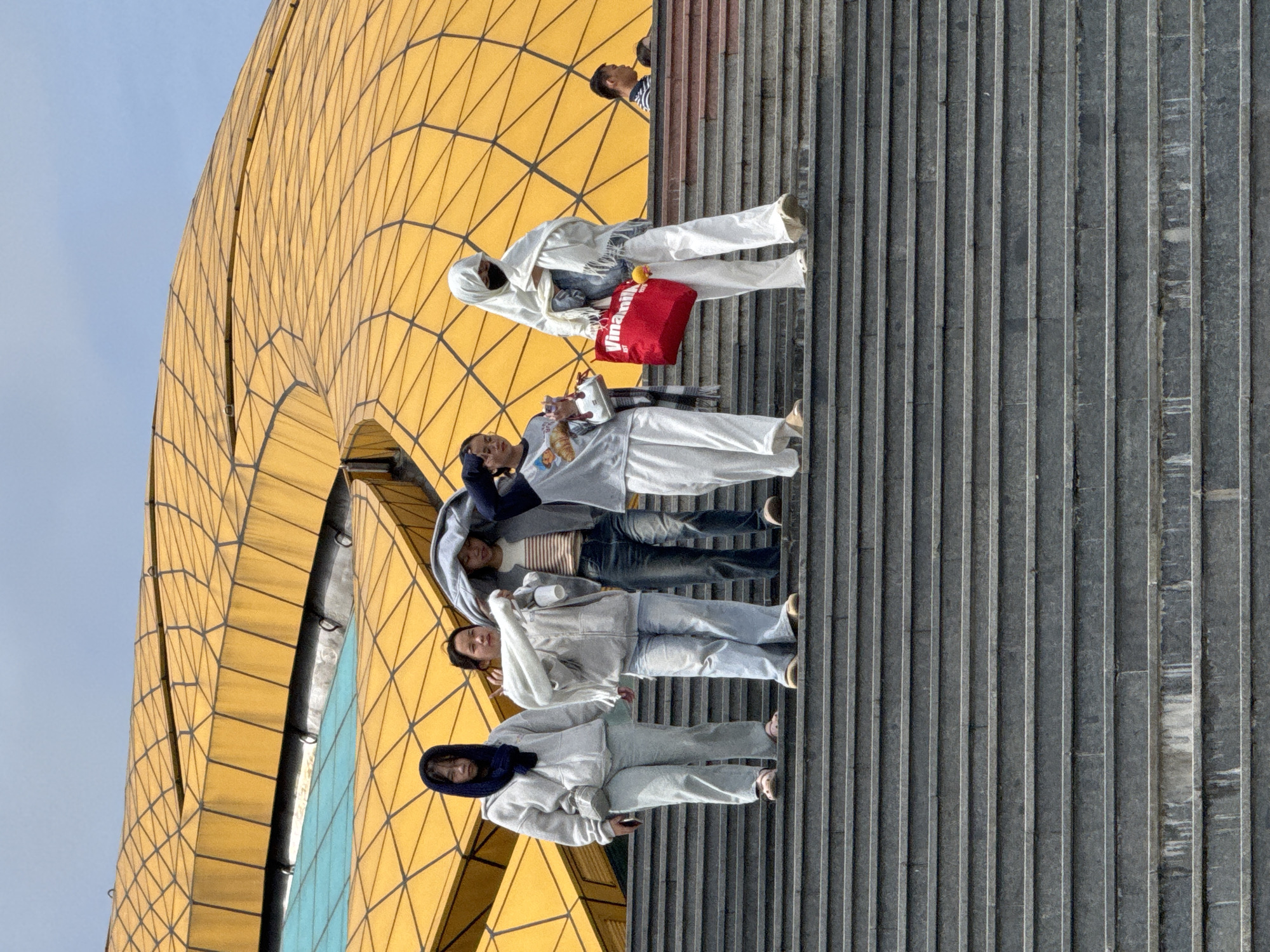 A group of five people are walking down a wide, grey stone staircase in front of a distinctive yellow, curved architectural structure under a hazy, overcast sky. The building features a grid-like pattern on its exterior and a section of turquoise-colored material. The individuals, appearing to be young women, are dressed in casual, light-colored clothing, with some wearing hoodies or headscarves. One person is carrying a bright red tote bag with "Vinamilk" printed on it. The scene is set outdoors, with the stairs leading downwards and the imposing structure forming the backdrop. It is likely a public space or a tourist attraction.