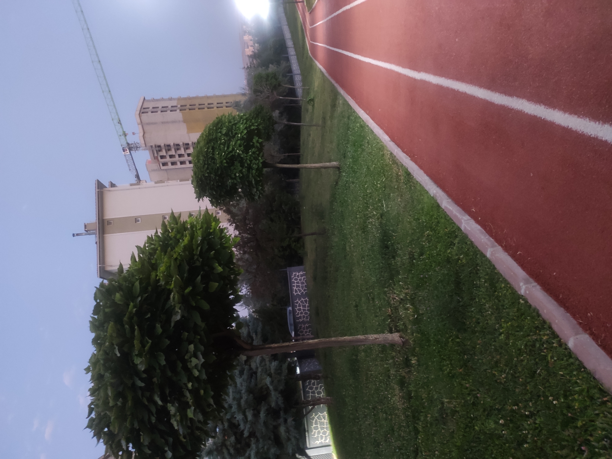 The image depicts an outdoor scene with a running track in the foreground. The track is red with white lane markers and is bordered by a curb and a grassy area. Several trees of varying sizes and types are present, some with full, lush canopies and others appearing more sparse. In the background, there are residential buildings and a construction crane, indicating ongoing development. The sky is a pale blue, suggesting it is either early morning or late evening. The overall environment appears to be a park or recreational area within a developed urban setting. Based on the provided location of Etimesgut, Turkey, this scene likely represents a public amenity within that city. There are no people visible in the image.