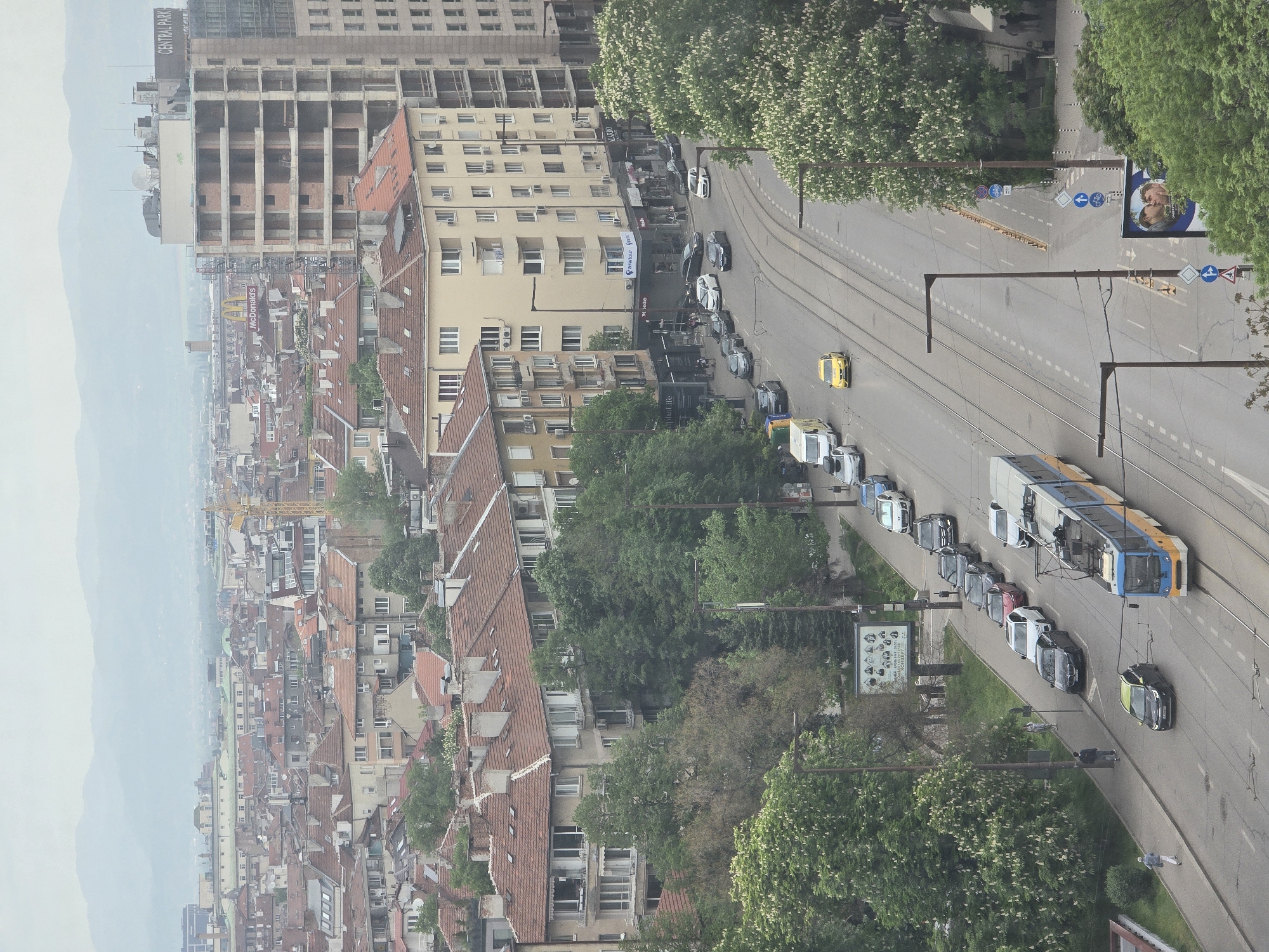 The image shows a street scene in Sofia, Bulgaria, taken from a high vantage point. The foreground is dominated by a wide street with tram tracks, along which a blue and yellow tram is traveling. Several cars are parked along the roadside. Street lamps and traffic signs are visible alongside the street. Adjacent to the road is a line of trees and greenery.
In the background, a dense urban landscape unfolds with buildings of varying heights and designs, many with red-tiled roofs. A taller building is visible in the distance, with a sign that appears to read "Central Park". Mountains are visible in the far background, shrouded in a light haze. The overall impression is that of a busy urban area in a European city, with a mix of old and modern architecture. The sky is overcast, suggesting it is daytime.