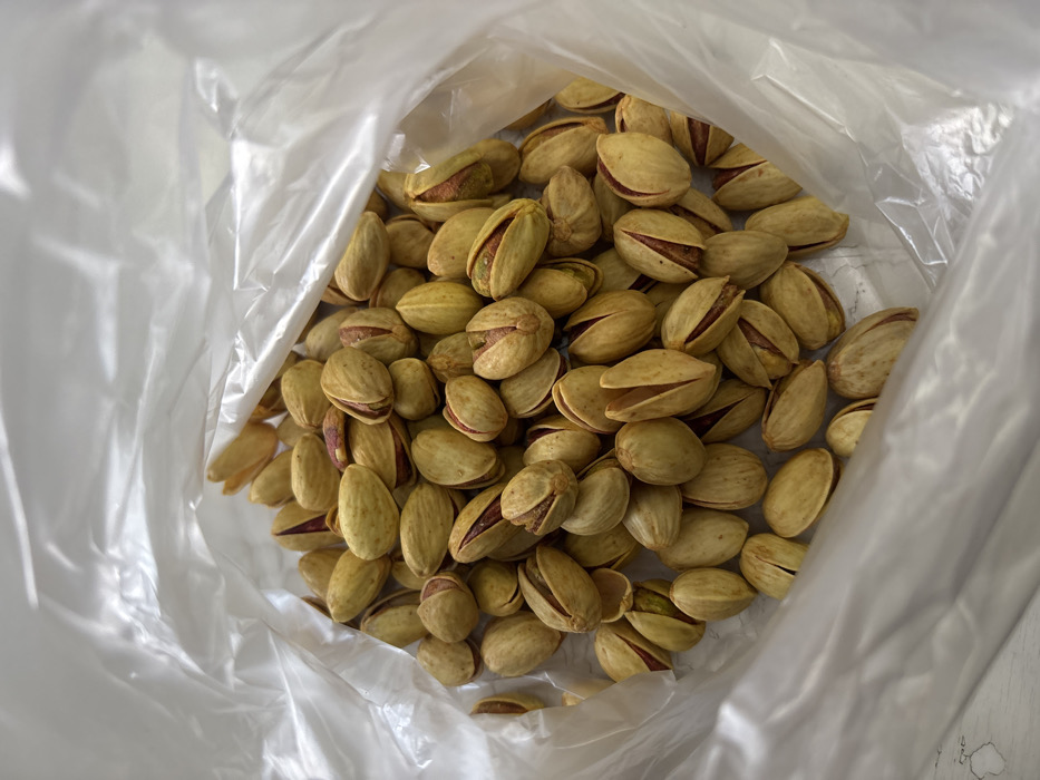 A clear, crinkled plastic bag is depicted from an elevated perspective, filled with numerous in-shell pistachios. The pistachios feature light tan to yellowish-green shells, with many naturally split open to reveal reddish-purple and green kernels. The interior of the bag is predominantly filled with these nuts, with slight variations in shell color and kernel visibility. The scene is located in Lehvaz, Armenia.
