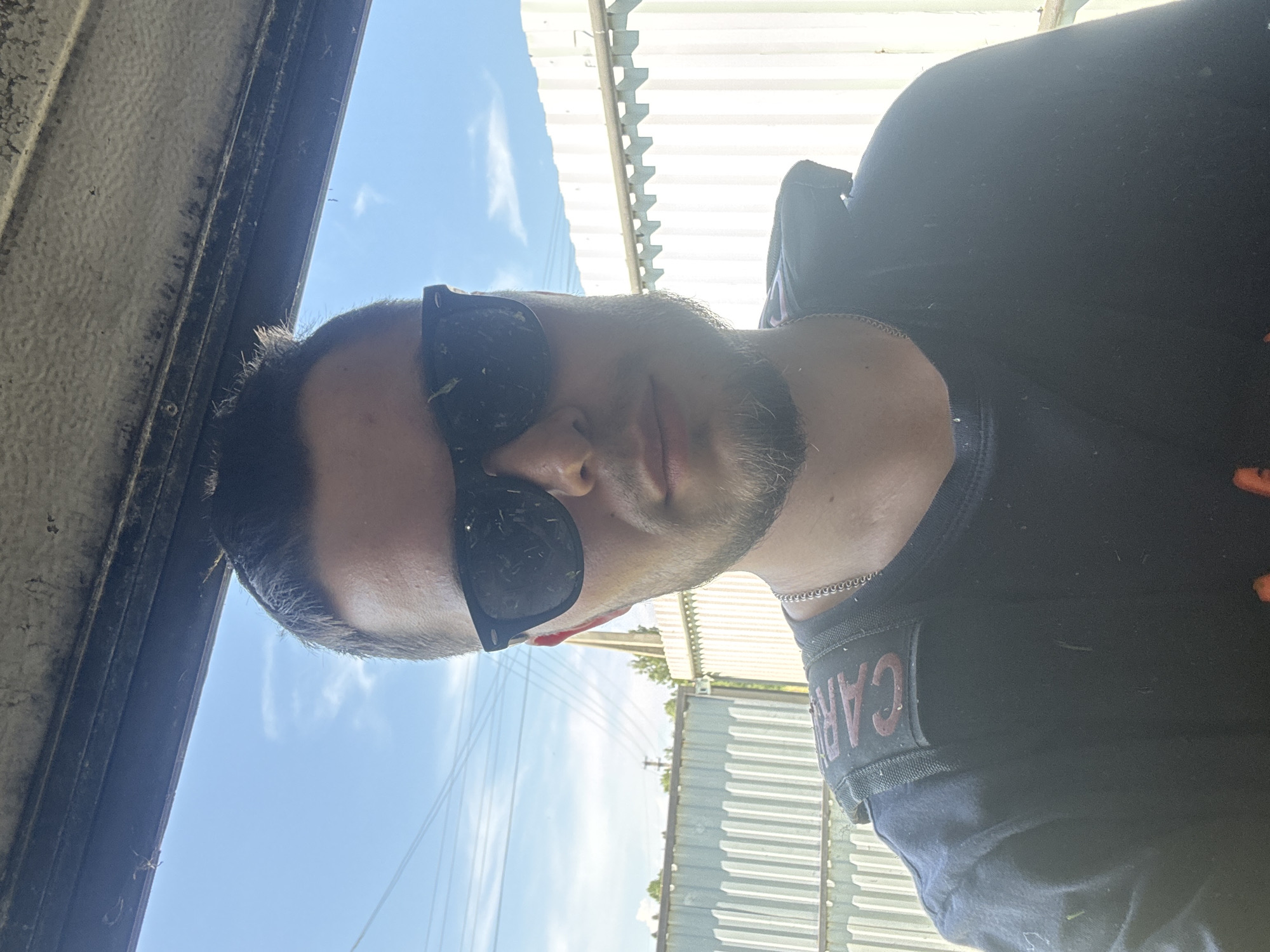 In a medium shot, a young adult male, seemingly in his early to mid-twenties, is positioned in the foreground, taking a selfie. He has short, dark hair styled upwards and is wearing dark-rimmed sunglasses that obscure his eyes. A light beard and mustache frame his face, and he has a thin silver chain visible around his neck. He is wearing a dark t-shirt, and a black strap with a red embroidered patch reading "CAR" is visible across his left shoulder. The background consists of a bright blue sky with scattered white clouds, and a corrugated metal fence runs horizontally behind him. A dark, weathered overhang is visible at the top of the frame. The overall lighting suggests a sunny day. The specific location within Borodulino, Russia, is not discernible from the visual information provided.