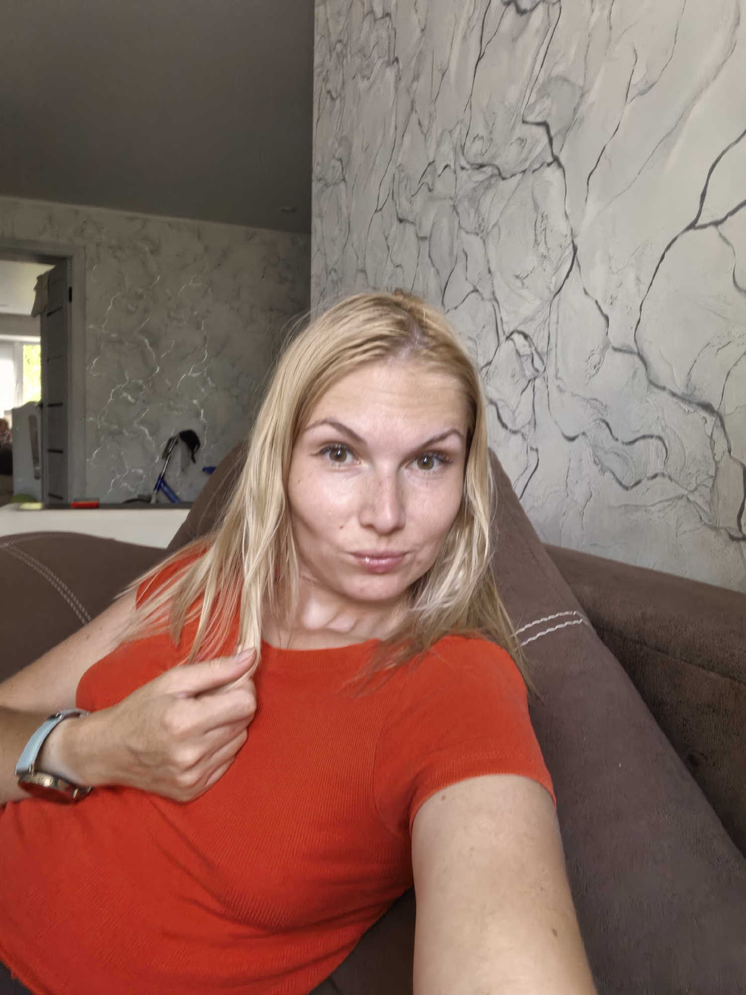 A woman with blonde hair, wearing an orange shirt and a watch, is sitting on a brown couch. She is holding a strand of her hair. The background shows a textured wall and a doorway leading to another room. The scene appears to be indoors. The image does not provide any information about the location in Sterlitamak, Russia.