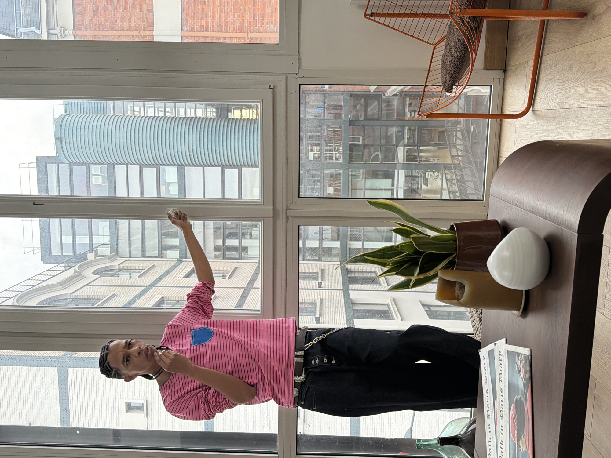 A full shot shows a person standing by a large window in an interior setting. The person is fair-skinned and dressed in a pink striped long-sleeve shirt with a blue patch, black pants, and a prominent silver belt chain. They are facing the window with their left arm raised and holding a small, unclear object. Their right hand is near their face.
The view outside the window shows multiple buildings, including a distinct cylindrical building with horizontal bands and other architectural structures, typical of an urban environment. The window frame is white, and the sky is overcast.
To the right of the person is a brown wooden table with a houseplant in a brown pot, a white sphere-like object, and a magazine titled "Walk in Paris Diary." An orange metal chair with a cushion is visible in the upper right corner.
The location is likely Saint-Denis, France, given the architectural style of the buildings and their configuration outside the window.