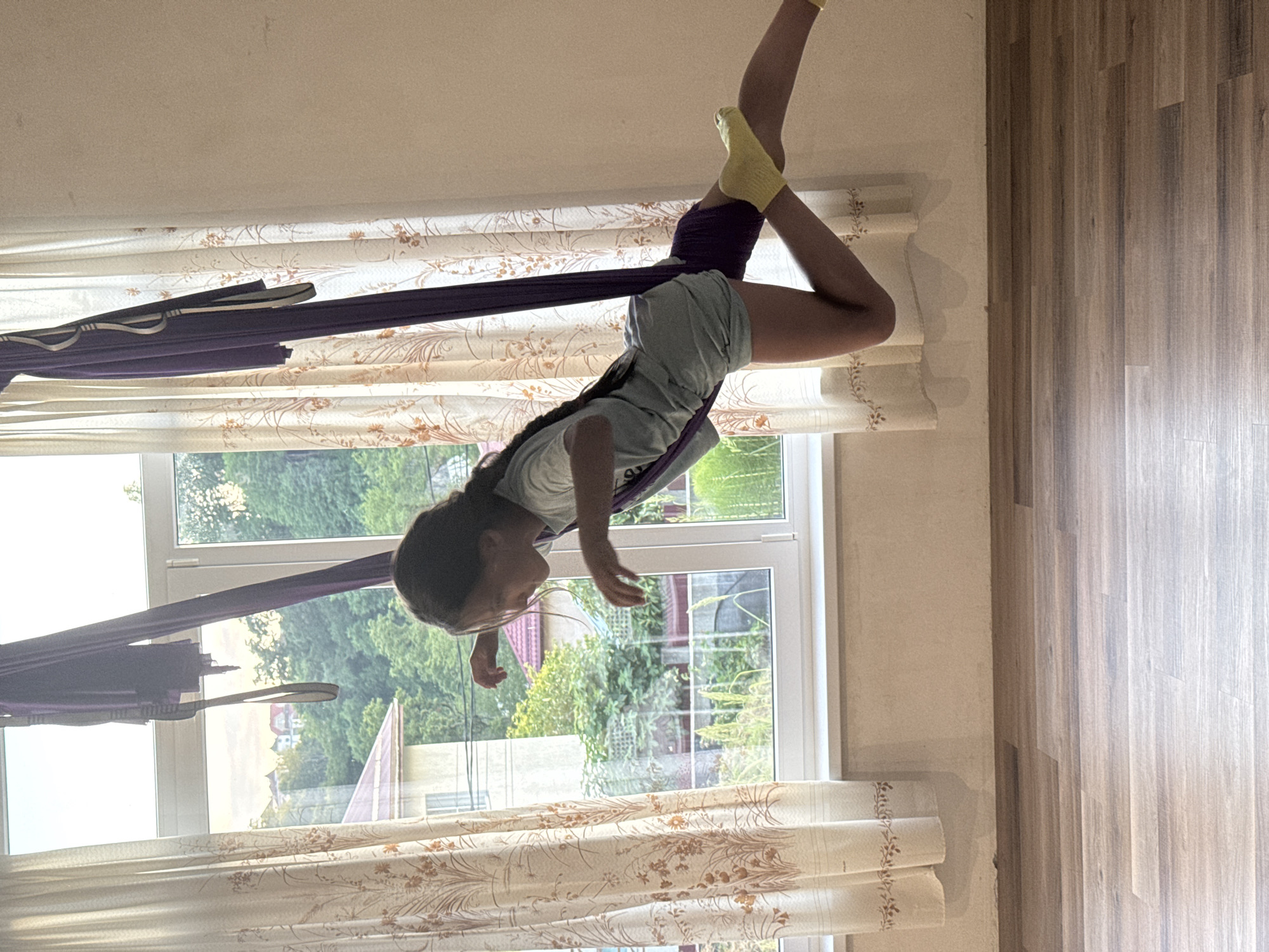 The image shows a young girl performing aerial yoga in a room with large windows. She is suspended in the air, supported by a dark purple aerial silk, with her legs extended and one foot pointed upwards. She is wearing a light blue t-shirt and grey shorts, and yellow socks. Her hair is dark and pulled back.
The room has floor-to-ceiling windows that look out onto a lush green outdoor scene, with trees and glimpses of houses. The light suggests it is daytime, possibly late afternoon given the warm glow. The curtains are white with a floral pattern. The floor is made of wood planks.
The girl appears focused on her movements, her body contorted into an athletic pose. There are no visible signs of text. The overall impression is one of active movement and peaceful focus in a bright, natural setting. The location is Verkhniy Yurt, Russia.