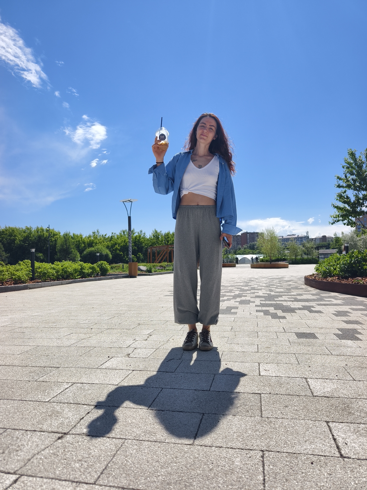 A young woman with long, wavy reddish-brown hair is captured in a low-angle shot, standing outdoors under a bright blue sky. She is looking directly at the camera with a neutral to slight smile. She wears a white cropped tank top, a loosely fitting light blue button-up shirt unbuttoned over it, and loose-fitting grey trousers. Her feet are clad in dark athletic shoes, and a tattoo is visible on her upper chest.
She holds up a clear plastic cup with a dark straw in her right hand, showcasing a light brown beverage, possibly iced coffee or tea. The cup has a circular white label with dark text that reads "BOBA SPOT".
The setting appears to be a modern park or pedestrian area in Tyumen, Russia. The ground is paved with light grey and dark grey rectangular tiles, forming a subtle pattern. Lush green trees and bushes line the edges of the paved area, with some dark lampposts visible among the greenery. In the background, distant buildings and structures can be seen under a partly cloudy sky, though most of the sky is clear blue.
The sun is high, casting a prominent, elongated shadow of the woman and the drink she holds onto the paved ground in front of her. The bright sunshine suggests it is midday or early afternoon on a clear, warm day. There are no other people immediately visible, contributing to a sense of peacefulness.