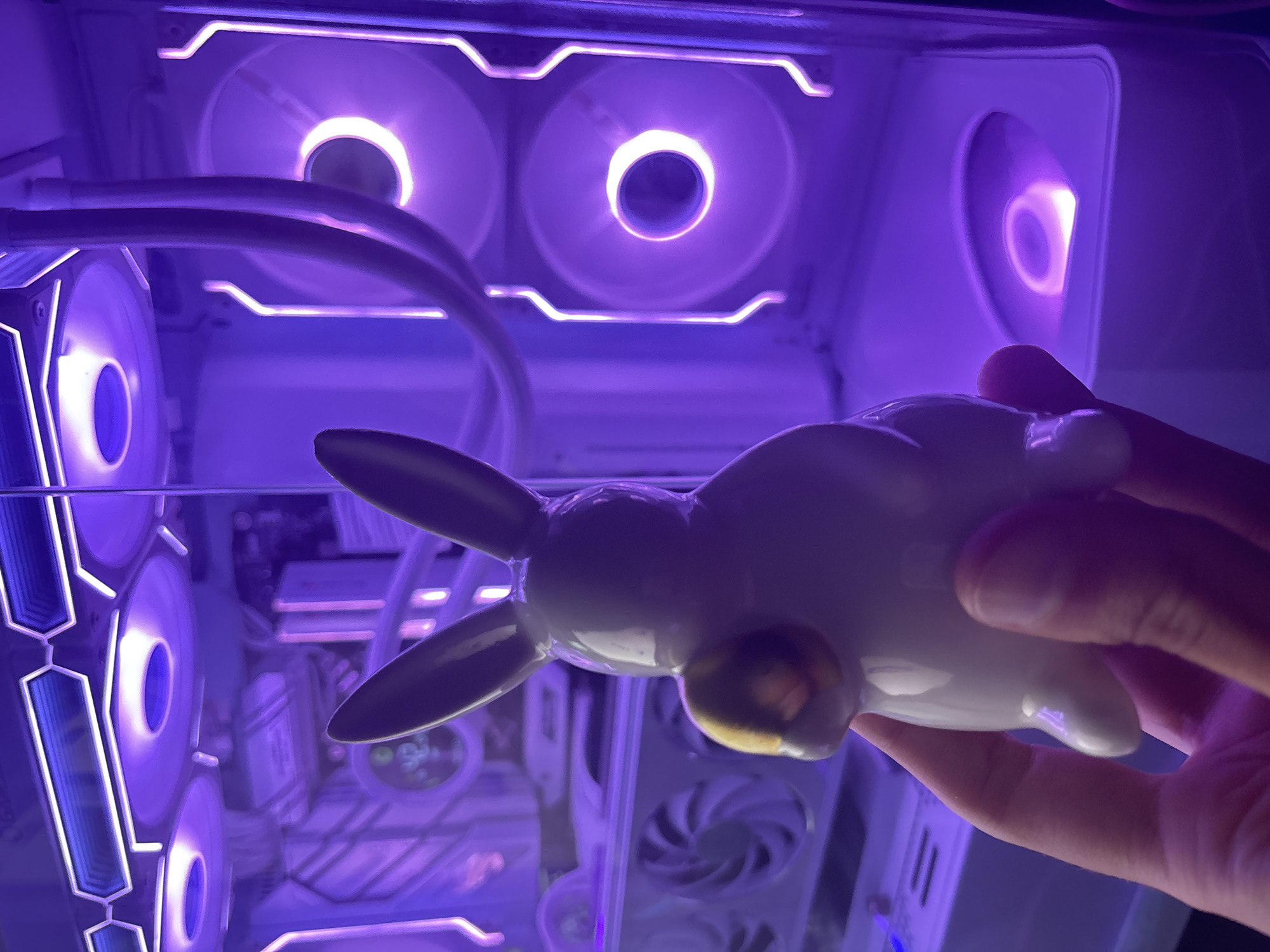 In this close-up, eye-level, indoor shot, a hand holds a white, ceramic bunny figurine with a gold sphere on its side, obscuring much of the figurine. The figurine is positioned in the foreground, with its back to the camera and ears pointed upwards. The bunny is held with the thumb and index finger of a person's right hand. The background reveals the interior of a white computer tower, illuminated by purple LED lighting. Several components are visible, including fans with glowing purple rings and other internal hardware. The overall lighting suggests a dimly lit environment with the primary illumination coming from the computer's lighting. The scene is not identifiable as being in any specific city or location.