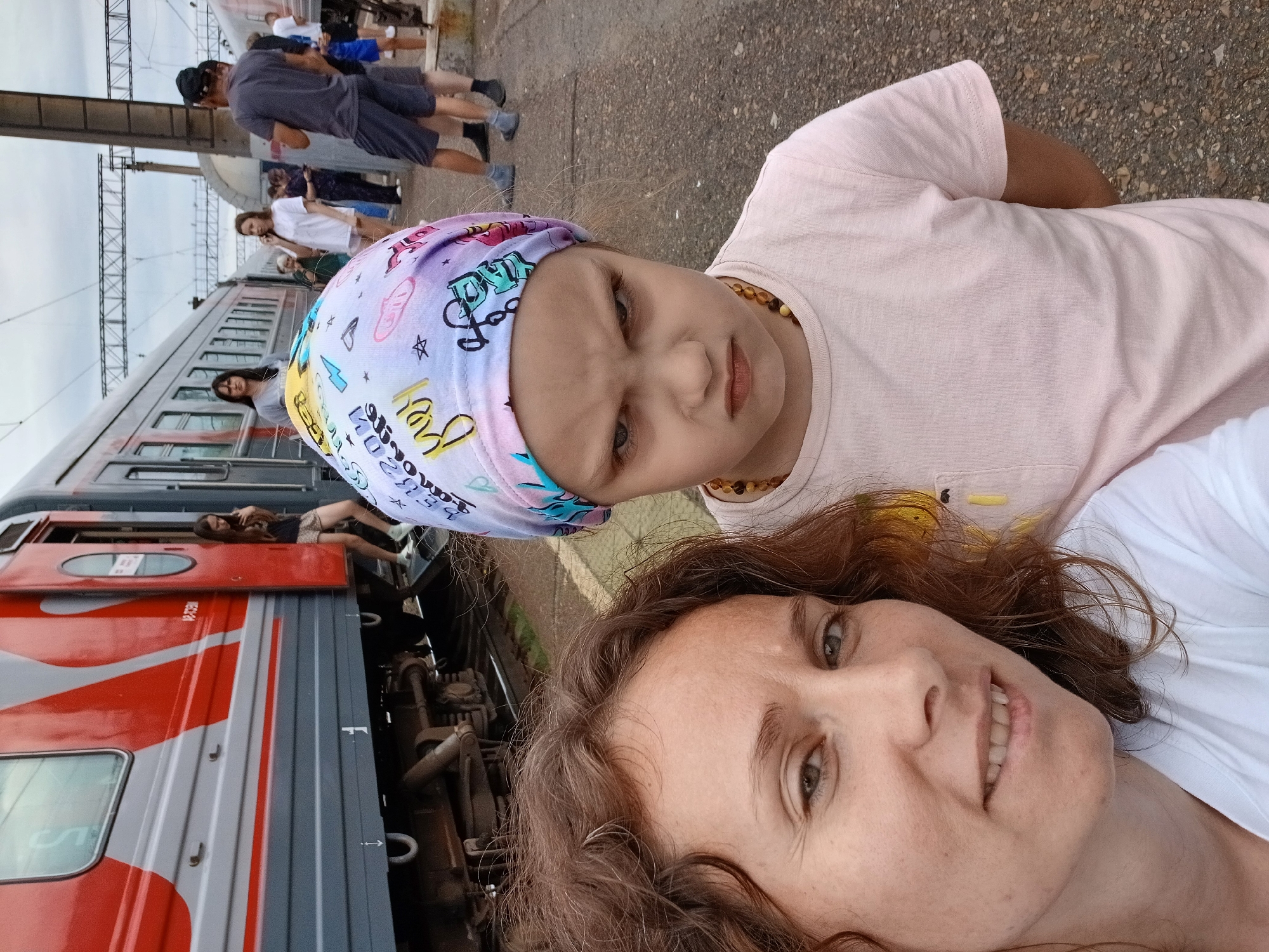The image captures a selfie of two people, an adult woman and a young girl, with a train and a train station platform in the background. The woman, on the left, has curly brown hair and is smiling. The young girl, on the right, is wearing a colorful headscarf and has an angry expression. She is wearing a pink t-shirt. In the background, people are seen standing on a train platform next to a red and gray train. The environment appears to be outdoors, with a cloudy sky overhead. The location is identified as the Kropachyovo, Russia train station.