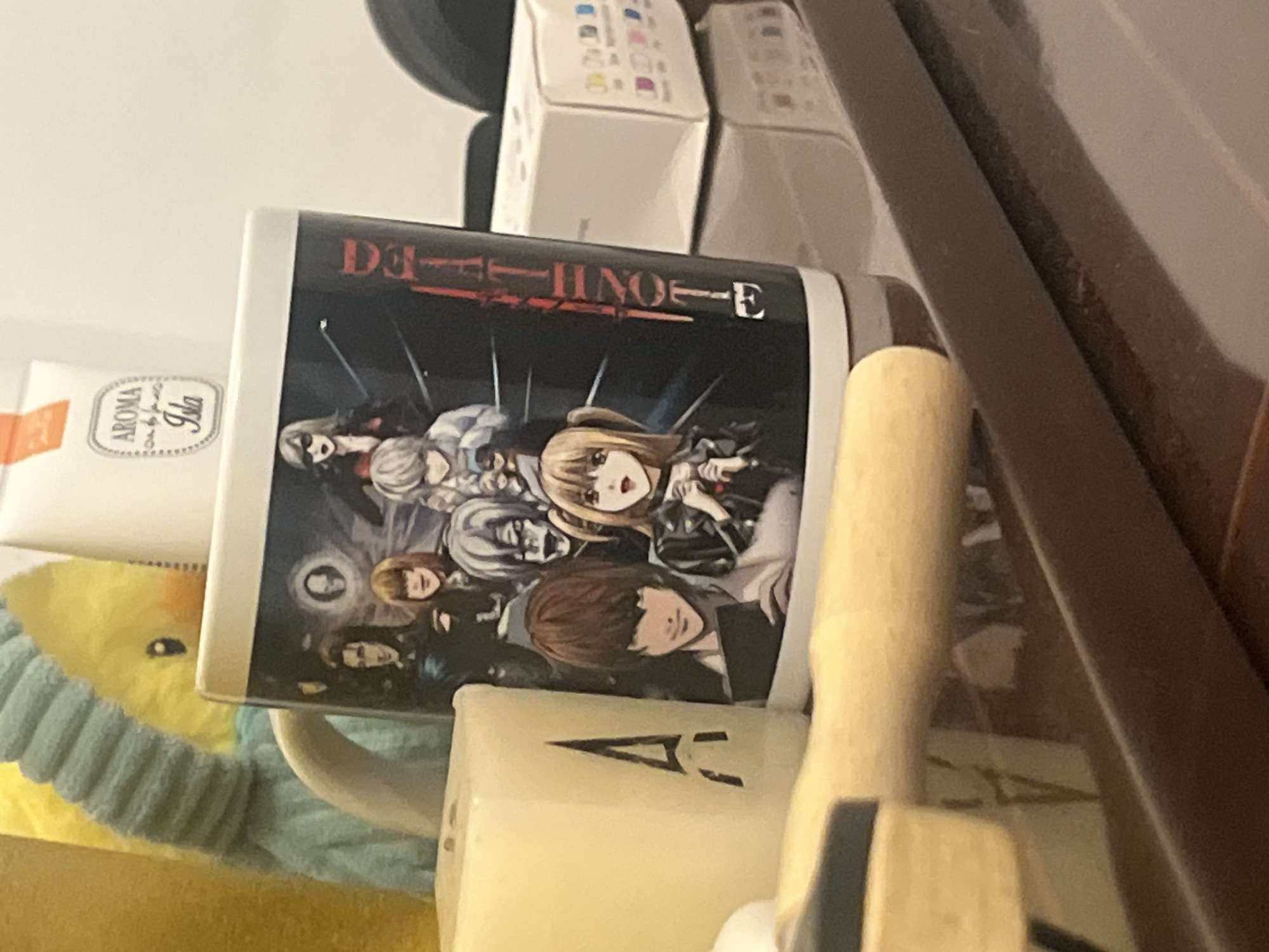 The image shows a Death Note mug, placed next to various items in an enclosed space. The mug has a black background with an image depicting characters from the anime series Death Note. Behind the mug are several white boxes, possibly containing ink cartridges. To the left of the mug, there is a yellow stuffed animal and a white box labeled "Aroma Isla". In the foreground, a wooden object is visible, obscuring some parts of the scene. The image does not provide enough information to locate it in Barcelona, Spain.