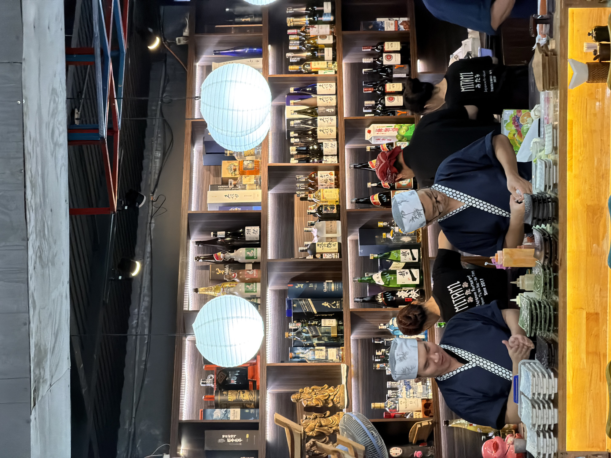 The image captures the interior of a Japanese restaurant or bar in Nha Trang Ward, Vietnam, presented with a clockwise 90-degree rotation. The scene, illuminated by artificial lighting, suggests evening or nighttime. In the foreground and mid-ground, two individuals, likely chefs, are seen working behind a long wooden counter. They wear blue uniforms with white patterned headbands. The person in the foreground is engaged in an activity at the counter, which is laden with small dishes, trays, and various condiments. Further back, another staff member in a black t-shirt is visible.
Behind the counter and extending across the back of the establishment are large wooden shelving units, richly stocked with an extensive collection of liquor bottles, including sake, wine, whiskey, and other spirits. Brands such as TALISKER, BLACK LABEL, Glenfiddich, ICHIKO, KUBOTA, Aberfeldy, Royal Salute, and Macallan are visible on various bottles and boxes. The shelves are internally lit by LED strips, highlighting the bottles. Two prominent white, spherical paper lanterns hang in front of these shelves, casting a soft glow.
On the far left, a textured grey wall and a decorative structure with red and blue geometric patterns are visible. A blue electric fan is partially seen in the lower foreground. Visible text includes "MITAMI", "みたみ", "Japanese Restaurant", "yen Thi Minh Khai-Nu Tray", and "Since 2017" on the black staff shirt. Other text on bottles or boxes includes "豪快" (Gōkai - Splendid/Dynamic), "直に" (Naoni - Directly), and "한우소주" (Hanwoo Soju - Korean beef soju). The overall atmosphere indicates a functional and aesthetically arranged dining space.