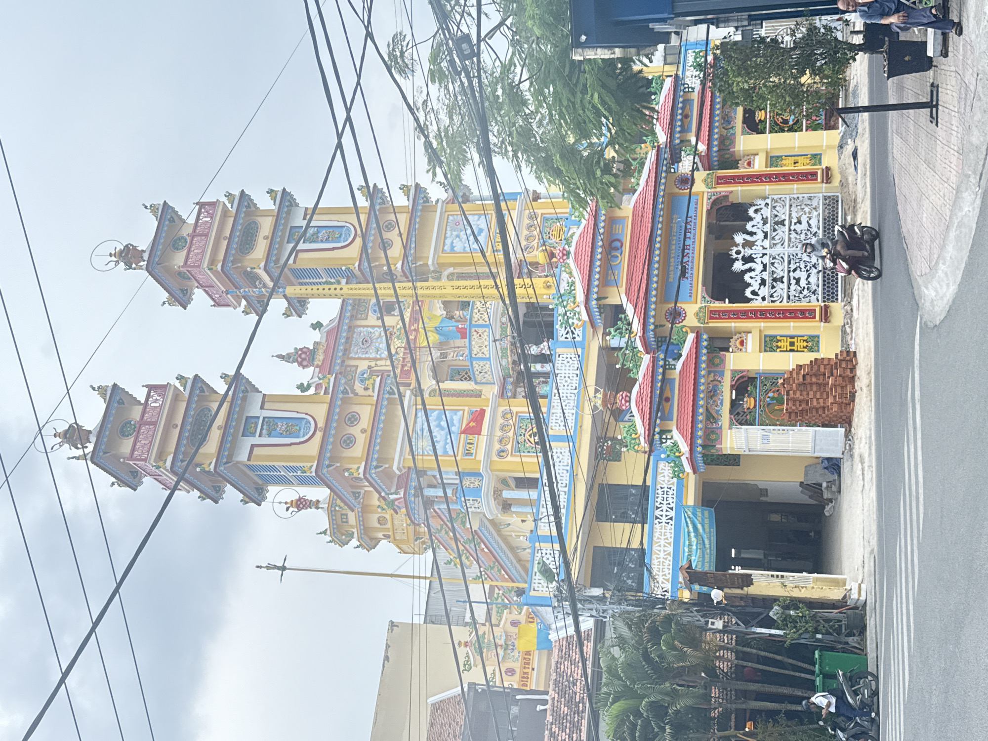The image depicts a colorful Buddhist temple in Nha Trang Ward, Vietnam. The temple has two tall, elaborate towers with intricate carvings, and a main building with a decorative entrance. Power lines crisscross in front of the building. A motorcyclist passes by. On the sidewalk to the right, a person sits at an outdoor cafe. A Vietnamese sign at the temple entrance reads "Dien Tho Di", "Loi Am" and "Thanh Thai". The sign also contains Chinese characters. There is construction equipment and stacked bricks near the building's main entrance. The sky is overcast.