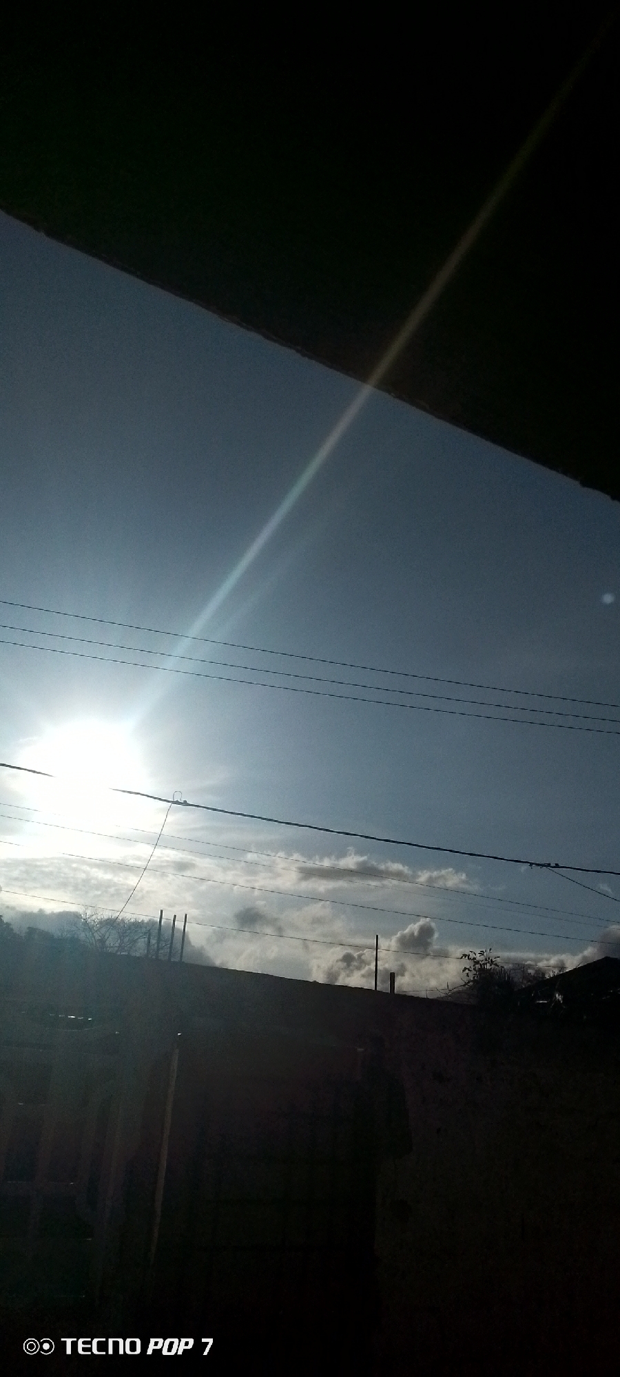 A view of a sky taken from an obscured foreground. The sky is mostly clear with some wispy clouds near the horizon line. A bright light source, presumably the sun, is shining from the left of the frame. Several power lines stretch across the sky. The location is Altagracia, Venezuela. The foreground is dark and indistinct. A structure, possibly a building, obscures the upper portion of the frame. There is no visible evidence of people or specific actions.