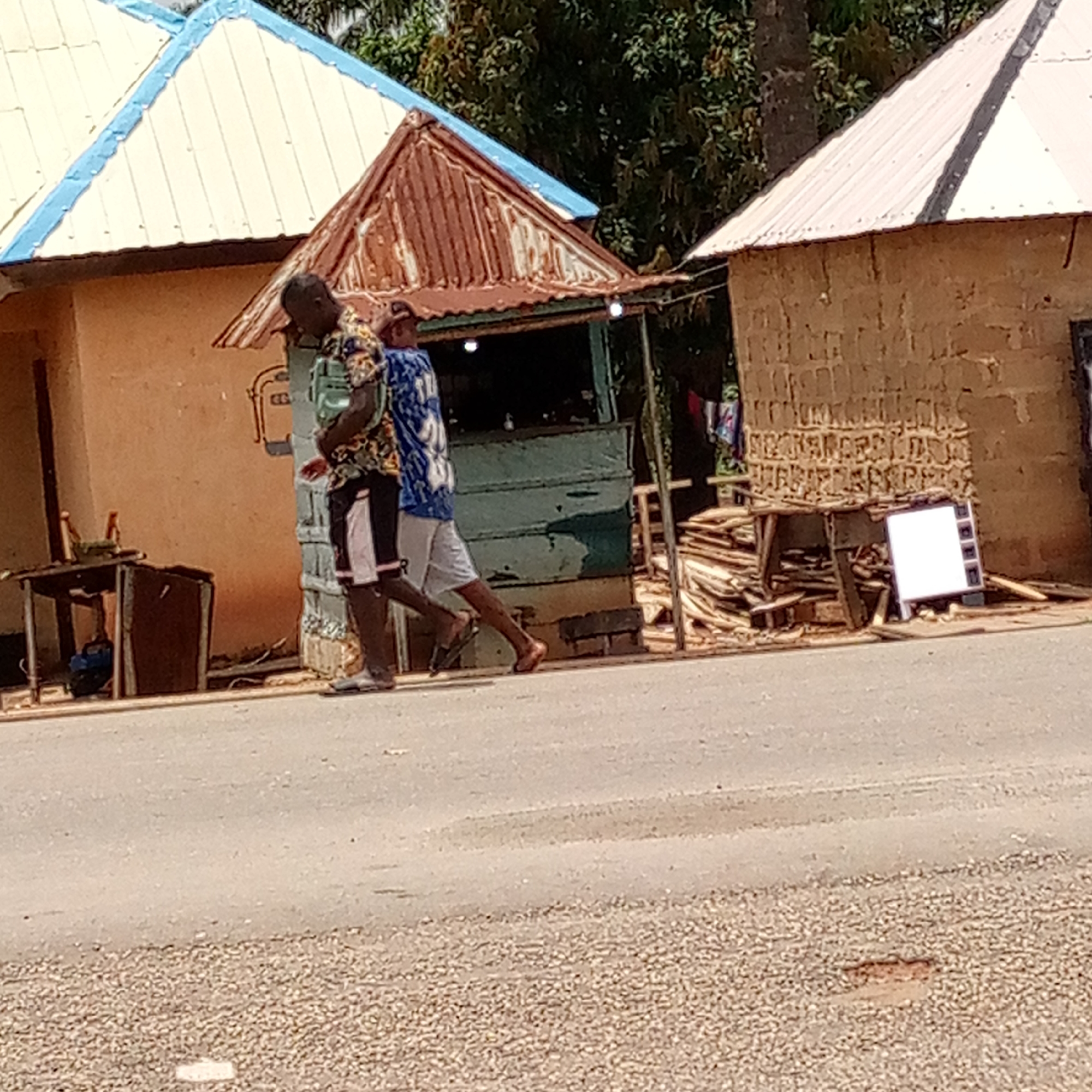 In Mararaba, Nigeria, a street scene captures two people walking along a paved road. The first individual wears a patterned shirt and dark shorts, while the second is clad in a blue and white shirt with light-colored shorts. Both are dark-skinned individuals. They walk past a series of small, rudimentary buildings. These structures appear to be basic dwellings or perhaps roadside businesses. One building has a rusted metal roof. Materials like wood scraps and other items are piled around one of the buildings. A blank, white sign is also visible. The surroundings seem to be a mix of residential and commercial activity. Trees are visible in the background.