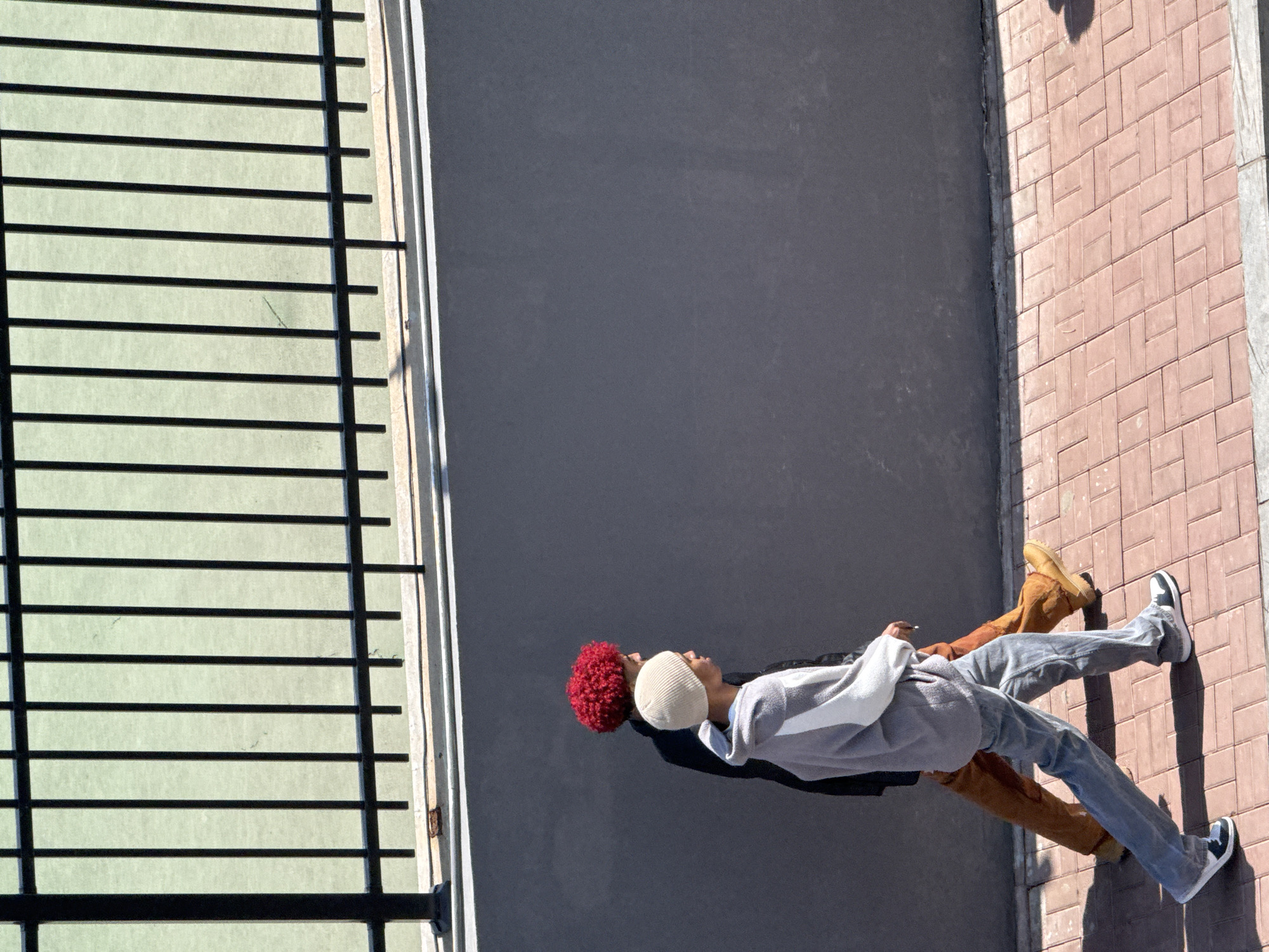 An overhead shot depicts two individuals lying side-by-side on a paved surface adjacent to what appears to be a building exterior in Zhongtun, China. The individual on the left has red, curly hair and is wearing a dark jacket. The individual on the right is wearing a cream-colored beanie, a light grey sweatshirt, and light blue jeans with black and white sneakers. This individual's legs overlap with the first individual's. The person closer to the bricked part of the ground is wearing timberland boots and brown pants. The paved surface is bordered by a brick walkway on one side and a building facade with horizontal slats on the other side. The ground is smooth and neutral-toned.