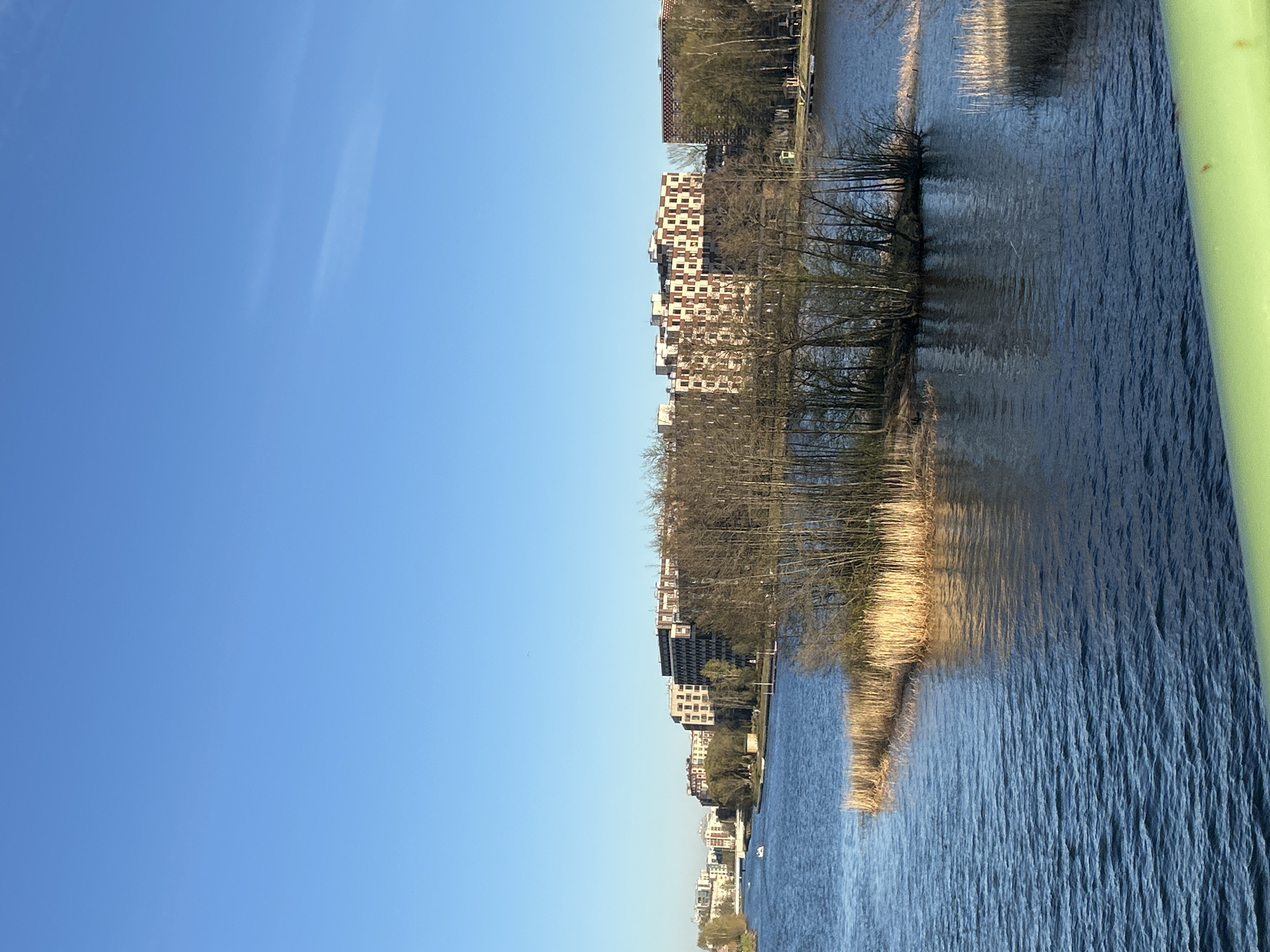 The image shows a view of the Neva River in Saint Petersburg, Russia. The sky is a clear blue. On the far side of the river, there is a row of modern residential buildings. Trees line the riverbank, their bare branches reflecting in the water. The water is dark and rippled. A green metal structure, likely a bridge railing, is visible in the right edge of the frame.