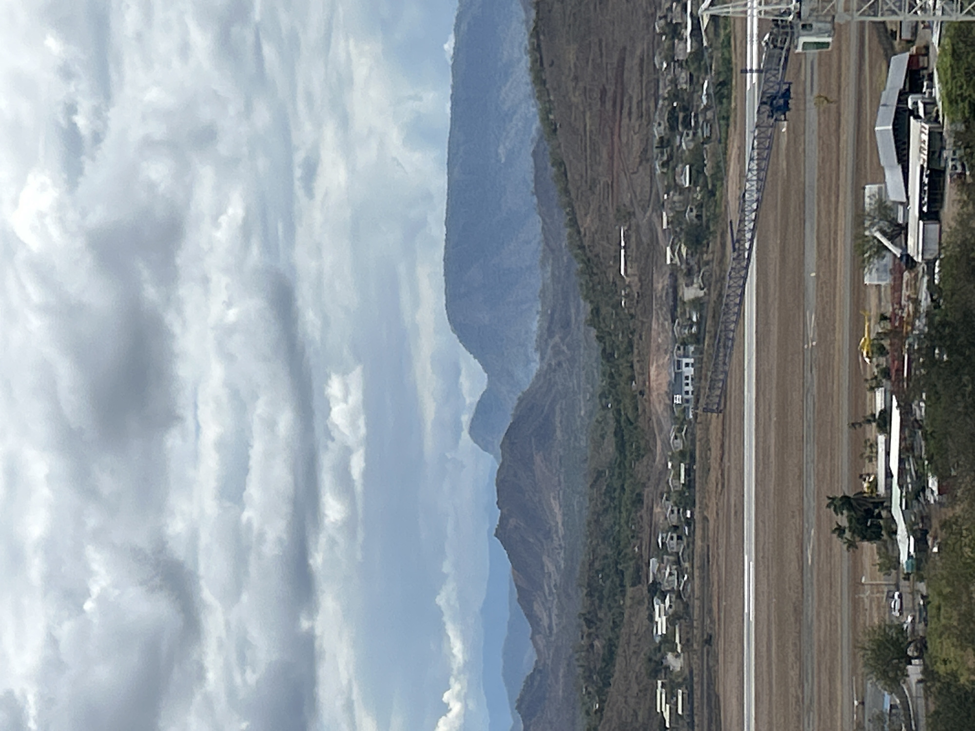 The image depicts a wide-angle view of a landscape with an airport runway in the foreground, likely in Port Moresby, Papua New Guinea. The sky is overcast with dramatic, grey clouds. Beyond the runway, there is a town or settlement with numerous houses and buildings spread across the terrain. Further in the distance, rugged, brown-toned hills and mountains rise, some with visible terracing suggesting agricultural use. A large construction crane is prominently visible on the right side of the frame, positioned near the airport. A small yellow helicopter is seen in flight near the runway. The overall scene suggests a developing area with ongoing construction and a significant natural landscape.
