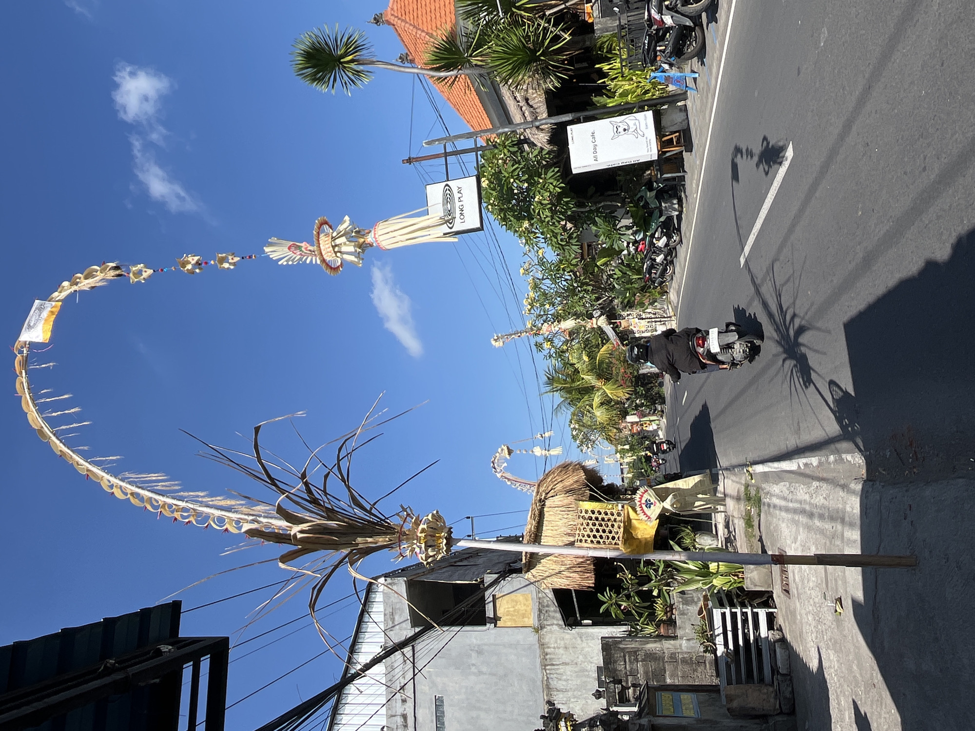 The image depicts a street scene in Mengwi, Indonesia, under a clear blue sky with scattered clouds. The road is paved and has a white line marking the center. A person is riding a scooter on the right side of the road, casting a long shadow. Along the left side of the road, there are buildings with various architectural styles, including some with thatched roofs and decorative structures made of natural materials, possibly representing cultural or religious elements. Prominently displayed are tall, arched decorations made of woven leaves and natural fibers, a common sight in Bali during certain festivals. One such decoration arches over the street, adorned with smaller ornaments. There are several parked motorbikes along the sidewalk. Signs are visible, including one for "LONG PLAY" and another for "All Day Cafe." Vegetation, including palm trees and flowering plants, is present, adding to the tropical atmosphere. The overall impression is one of a vibrant street in a Southeast Asian town, likely Bali, given the distinctive decorations.