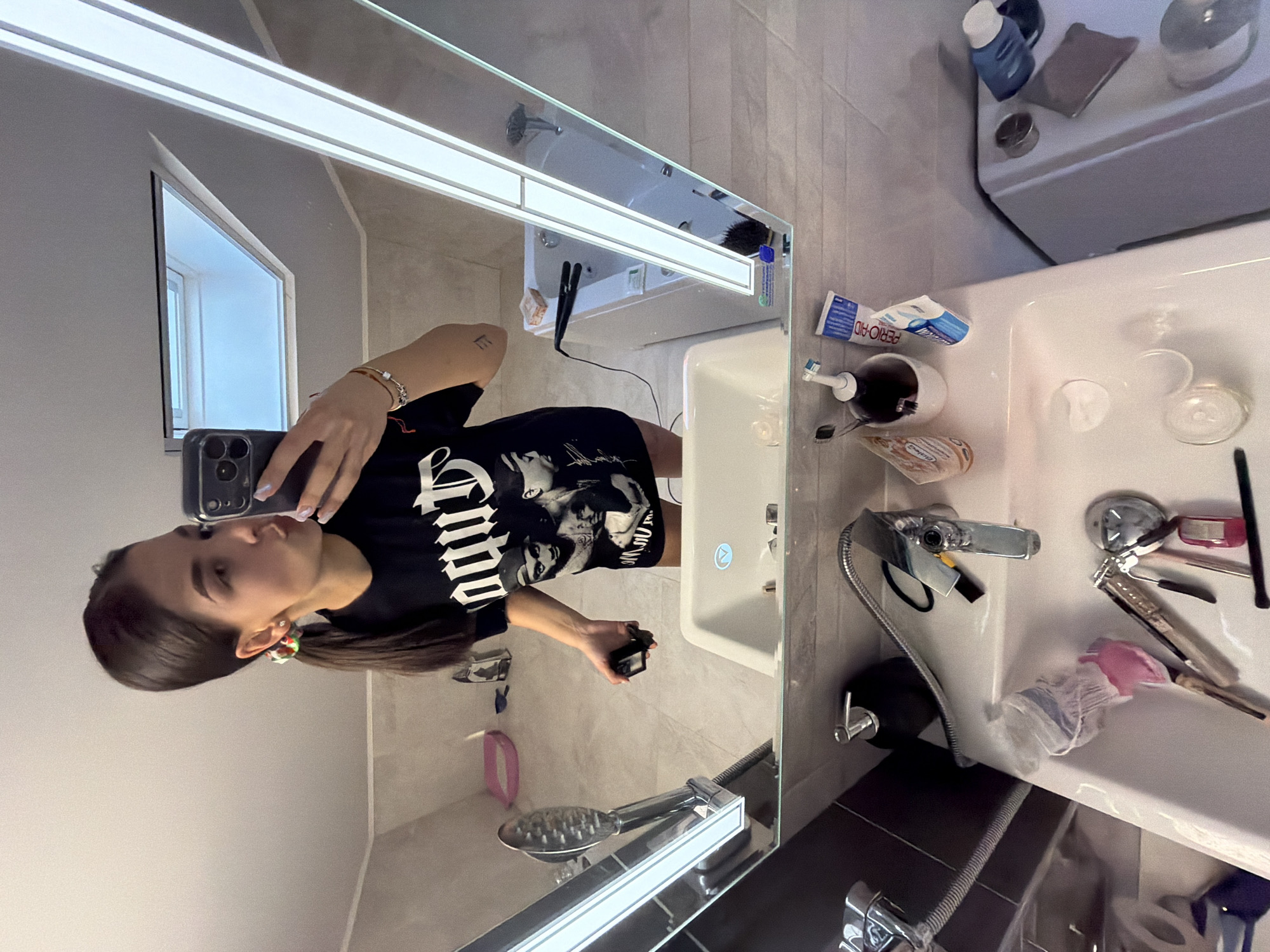 A selfie taken in a bathroom. A woman with dark hair pulled back into a ponytail is holding a phone in front of a mirror. She is wearing a black t-shirt. The bathroom features a sink with toiletries and cosmetics scattered around it, a mirrored cabinet, and a shower. The image does not provide enough information to identify the location in Gabrovo, Bulgaria.