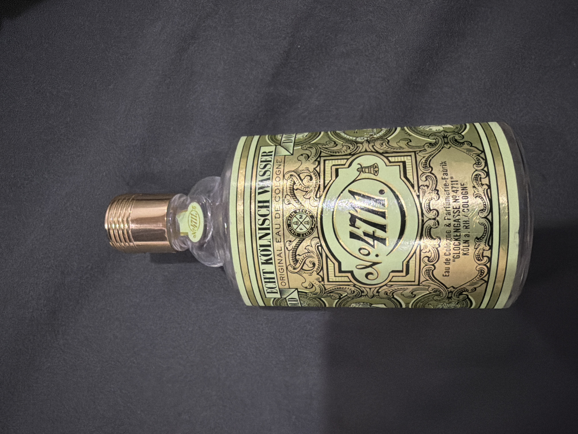 The image shows a bottle of 4711 Echt Kölnisch Wasser (Original Eau de Cologne) lying horizontally on a dark, textured surface. The bottle has a clear body and a gold-colored cap. The label is a pale green and gold design, prominently featuring the "№ 4711" logo. Text on the label includes "ECHT KÖLNISCH WASSER", "ORIGINAL EAU DE COLOGNE", and information about the cologne's manufacturer: "Eau de Cologne & Parfümerie-Fabrik 'GLOCKENGASSE N№ 4711' KÖLN a. RH./COLOGNE".