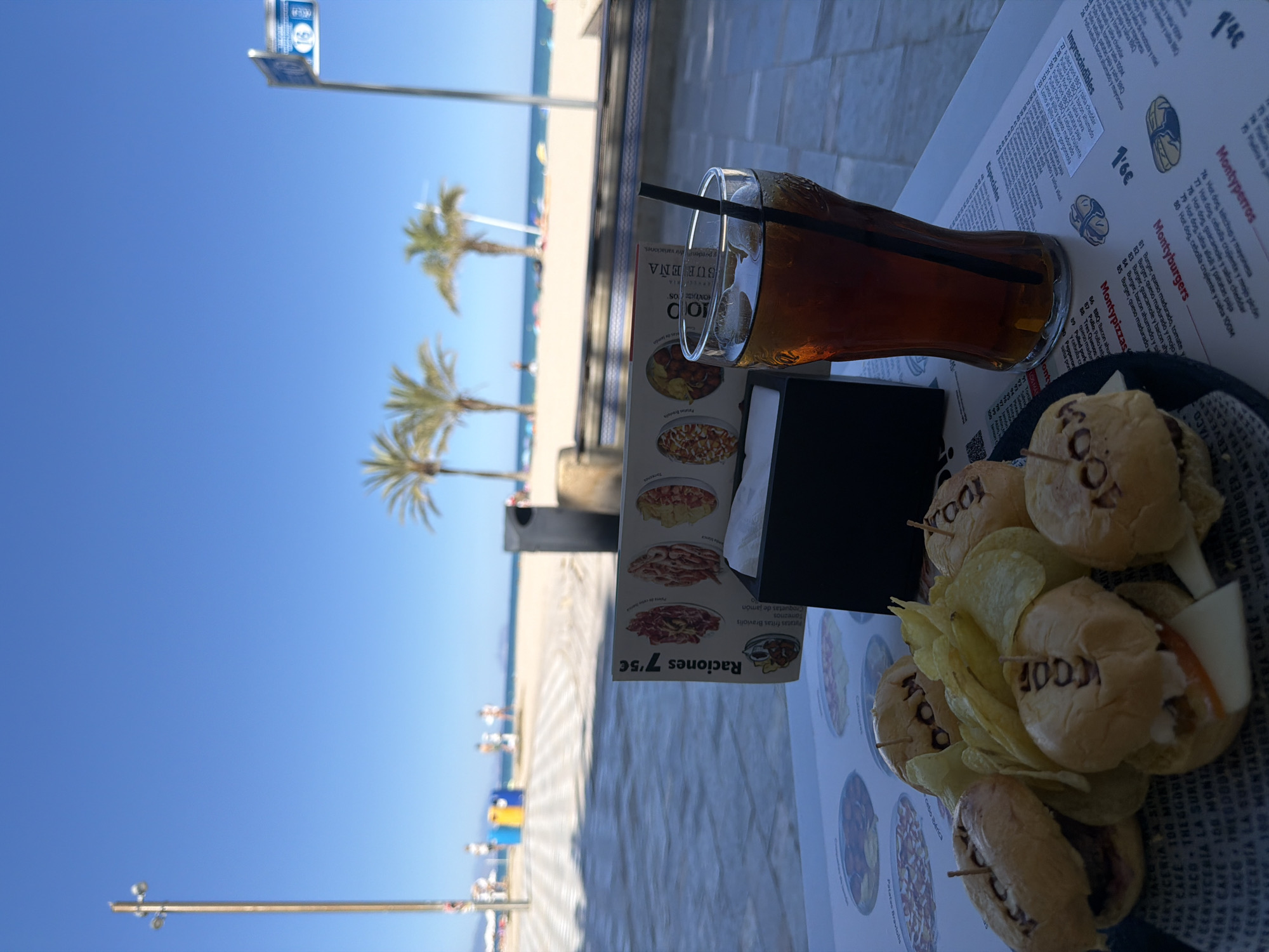 The image shows a table at an outdoor restaurant, likely near the beach in Alicante, Spain. On the table is a glass of iced tea with a straw, a black napkin holder, a food menu, and a plate of small sandwiches labeled "100." The plate also contains potato chips. The beach and the blue sky are visible in the background. Palm trees line the beach. The picture is taken at an angle.