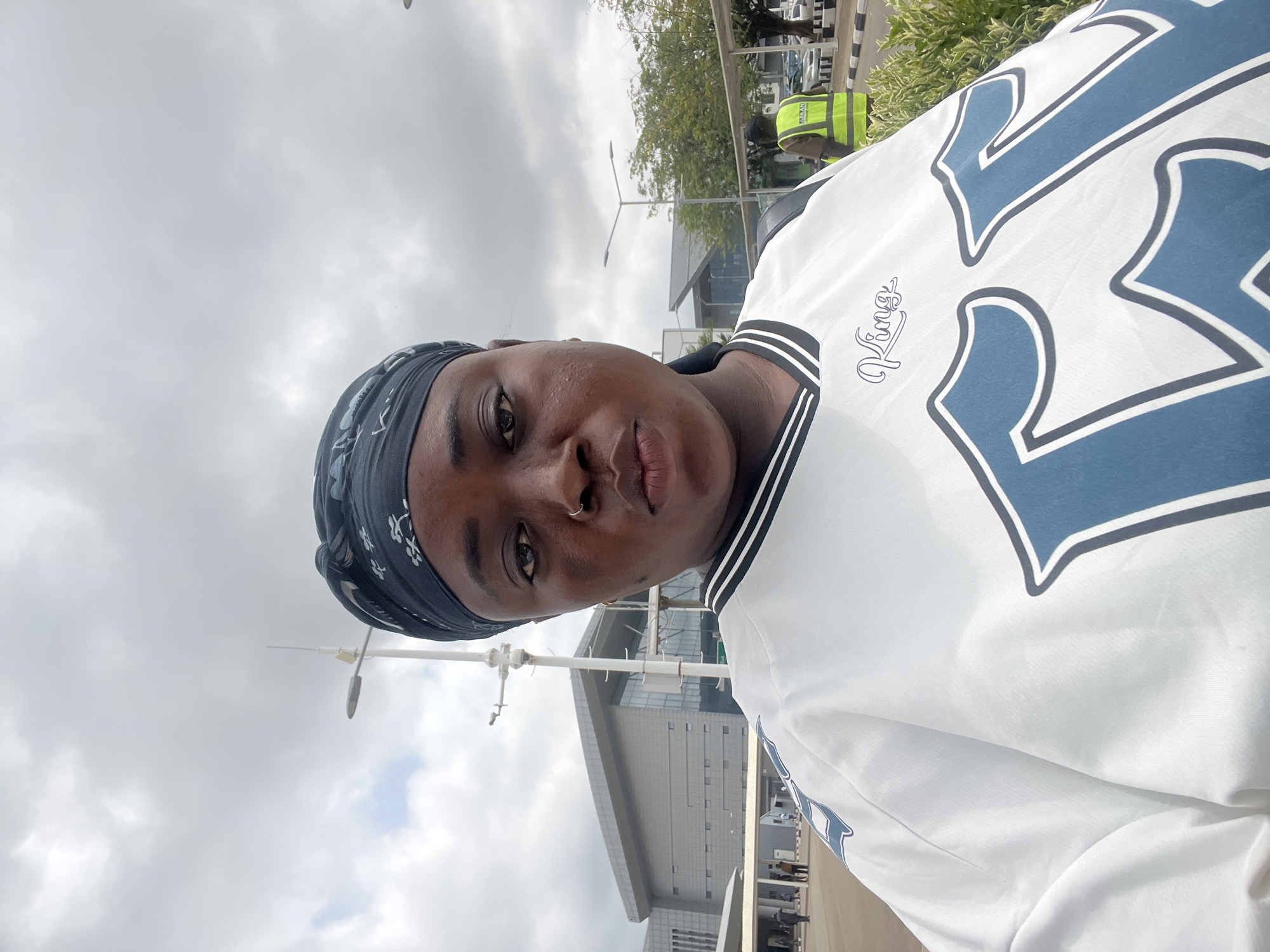 This image is a low-angle selfie, rotated 90 degrees clockwise, featuring a person with dark skin. The subject looks directly at the camera with a neutral expression. They are wearing a dark, patterned headscarf and a white short-sleeved top, possibly a sports jersey, with black and white striped trim. The shirt prominently displays large blue numbers, likely "23," outlined in black, and a smaller cursive text reading "Kings" on the upper left chest area. A small gold hoop nose piercing is visible.
The background shows an outdoor setting under a cloudy, overcast sky, indicating daytime. Modern, multi-story buildings with grey and glass facades are visible, suggesting a large urban complex or an airport terminal. Green trees, bushes, and several utility poles or streetlights are also present. In the mid-ground, another person wearing a bright yellow/lime green high-visibility vest can be seen, likely a worker or security personnel. The scene suggests a public space, consistent with an airport or large facility in Gwaska, Nigeria.