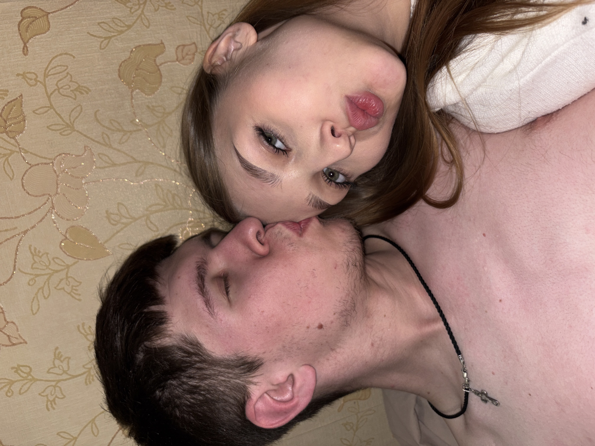 An intimate indoor portrait features two young adults, a female and a male, positioned closely together in Chaikovsky, Russia. They appear to be lying on a bed or sofa, against a light-colored background adorned with a subtle floral and leaf pattern in beige and golden hues.
The female is on her back, looking directly at the camera with her head slightly tilted. She has long brown hair, expressive green eyes, and distinct eyebrows. Her full lips are pursed in a pout, conveying a playful or affectionate expression. She wears a white top or sweater, parts of which are visible on her upper body.
Beside her, the male lies on his side, turned towards her with his eyes closed and lips slightly parted, suggesting he is either about to kiss her or is in the act of kissing her. He has dark, short hair and visible stubble on his face. Around his neck, he wears a black braided cord necklace secured with a silver clasp and featuring a small, silver cross-shaped pendant.
The scene captures a moment of close connection and affection, emphasizing their intimate interaction in a private setting. No text is visible in the image.
