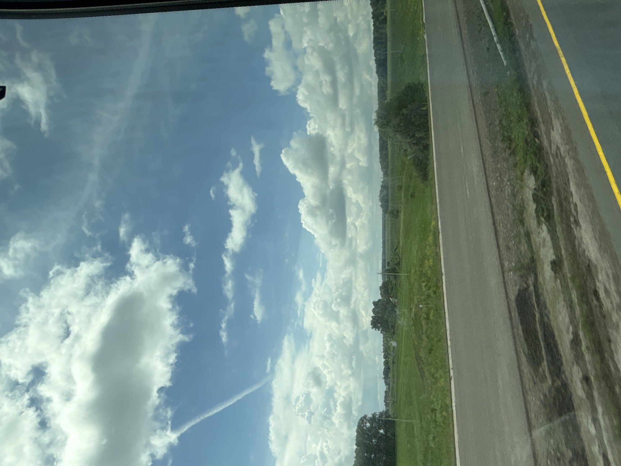The image depicts a rural landscape viewed from inside a vehicle, likely a car or bus, given the perspective through a window. The scene showcases a vast expanse of green fields under a bright blue sky dotted with fluffy white clouds and a thin contrail from an aircraft. A paved road with a yellow line runs horizontally across the foreground. The background features scattered trees and vegetation, suggesting a natural, undeveloped area. The location is identified as Taimurazino, Russia. There are no people or animals visible in the image. The overall impression is one of a serene, open, and natural environment on a clear day.