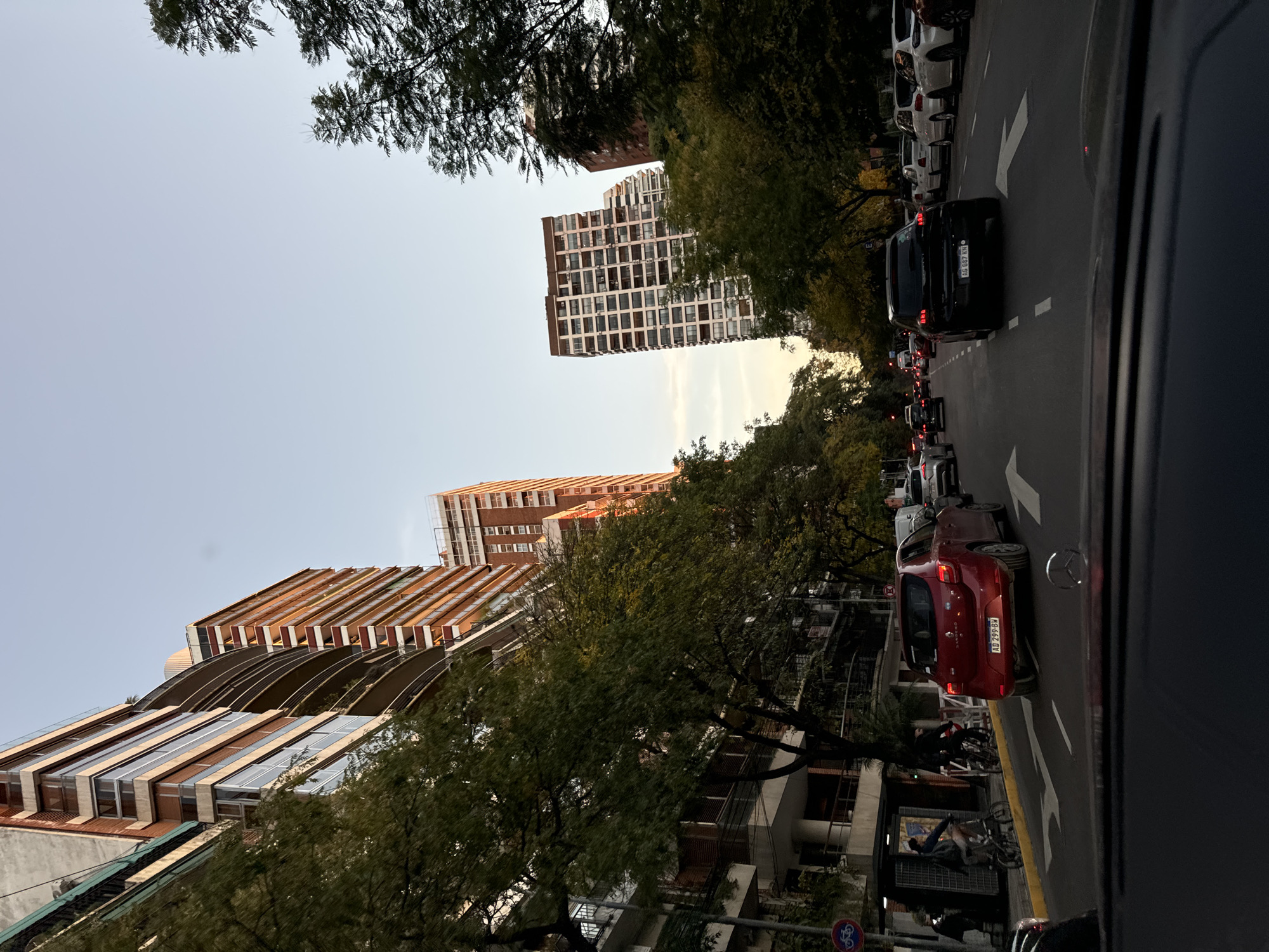 The image shows a street scene in Buenos Aires, Argentina, taken from inside a vehicle. The road is lined with tall buildings and trees. There are several vehicles in a traffic jam, including a red car in the foreground with the license plate "AD 299 BH". The buildings are a mix of older, traditional architecture and modern high-rises. The sky is overcast. On the sidewalk, there are people and bicycles. The scene captures a typical urban environment in Buenos Aires.
