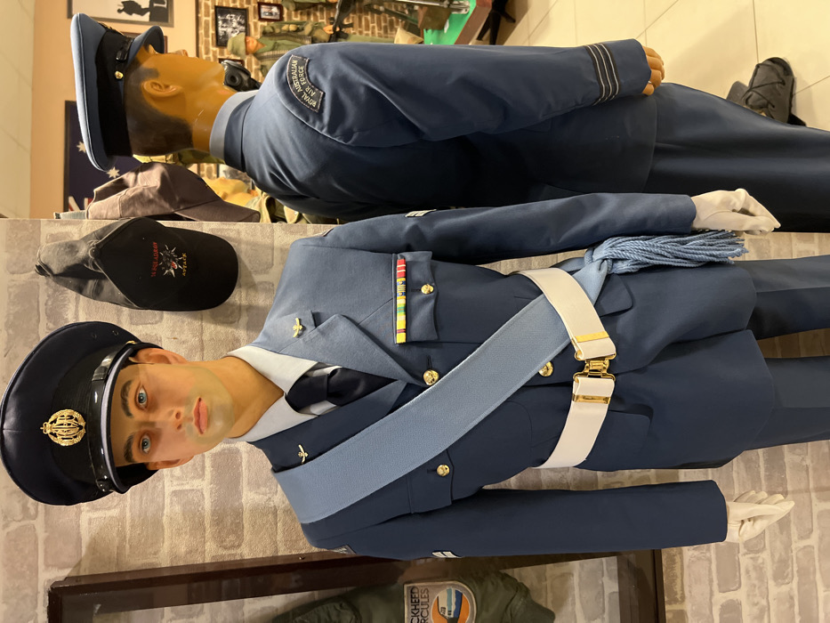Two mannequins are dressed in blue Royal Air Force uniforms. One stands taller, with a darker blue coat and hat displaying the RAF logo. The second mannequin wears a lighter blue uniform, complete with a dress cord, belt, and pilot wings. A black cap and khaki hat are visible nearby, along with a brick-patterned backdrop and other military-themed items.