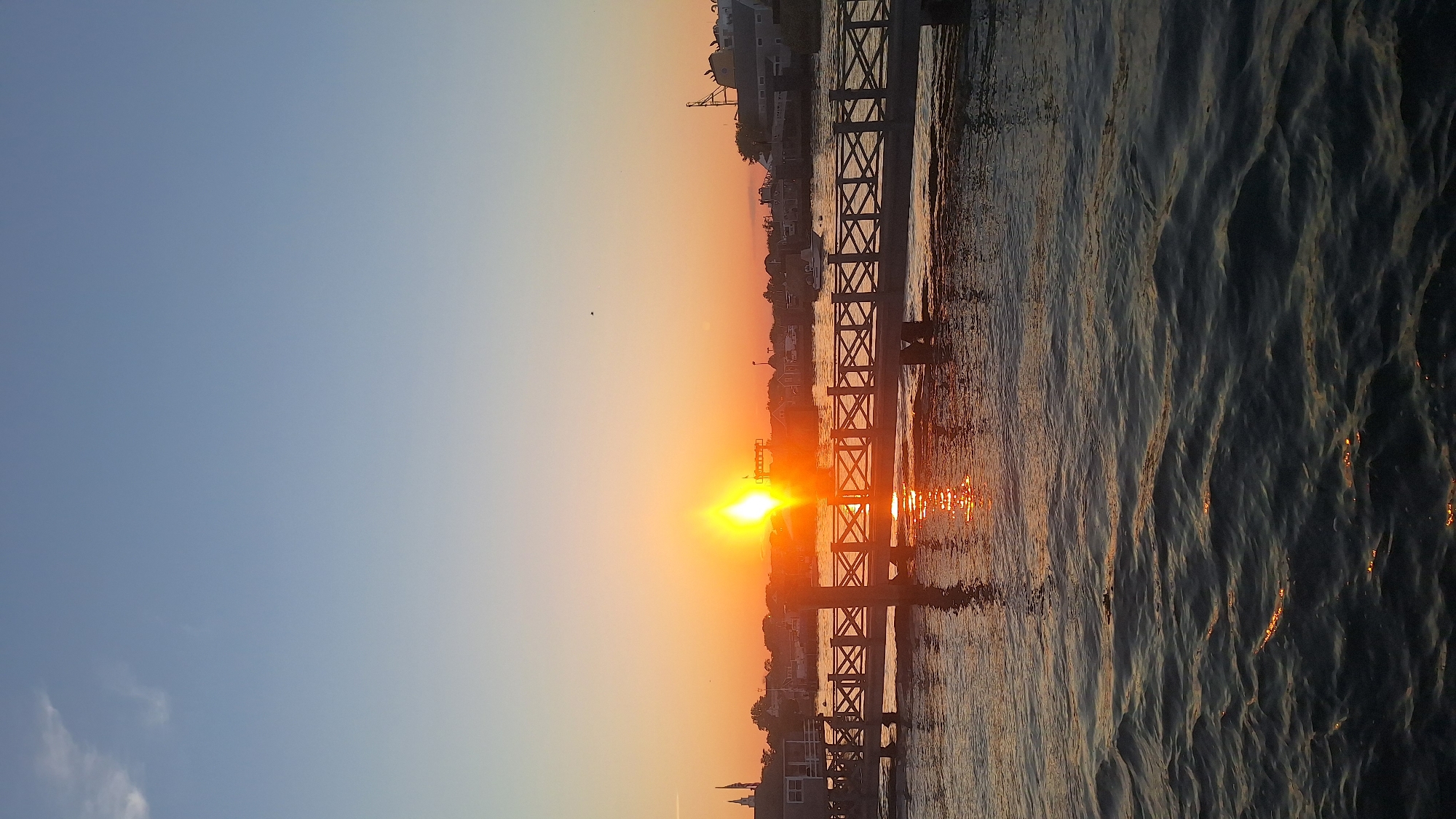 A sunset scene unfolds over the water in Nantucket, Massachusetts, USA. The sun, a brilliant orb, is low in the sky, casting a warm, orange glow across the horizon. Its rays are reflected on the rippling surface of the water, creating a shimmering path. A wooden boardwalk, with decorative cross-patterned railings, stretches across the foreground, supported by sturdy wooden pilings. Beyond the boardwalk, a collection of buildings, likely houses and businesses, cluster along the shoreline. Some appear to be commercial structures, possibly shops or restaurants, with details like awnings and signage visible. Boats are docked in the harbor, some with masts and rigging suggesting they are sailboats. A single small boat with a canopy is visible in the mid-ground. The sky above transitions from a pale blue at the top to a soft orange and pink hue near the horizon, indicative of the setting sun. A few wispy clouds drift in the upper portion of the sky. The overall impression is one of a tranquil coastal evening.