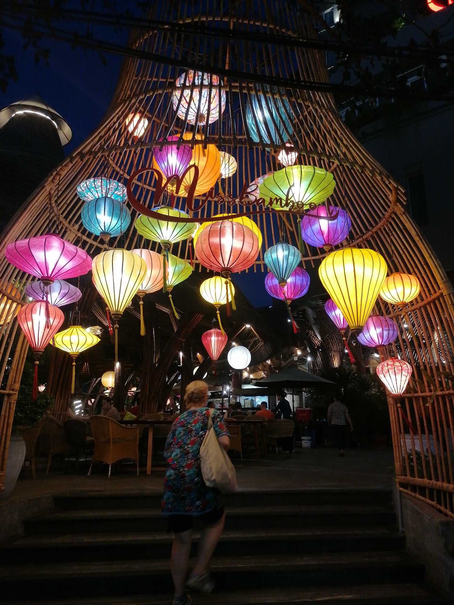The image shows an outdoor scene in Nha Trang Ward, Vietnam, at night. A tall, bamboo structure arches over a set of steps, adorned with numerous colorful lanterns of various shapes and sizes. The lanterns are illuminated, casting light on the steps and the surrounding area. A woman with blonde hair, wearing a floral-patterned top, dark shorts, and carrying a beige bag, is walking up the steps. The steps lead into an open area with tables and chairs, suggesting a restaurant or cafe. Other people can be seen seated at the tables or walking in the background. The sky is dark, and buildings are visible in the background, with some lights illuminating them. The overall atmosphere is festive and inviting.