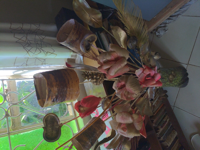 A bouquet of artificial flowers, consisting of gold and red silk blossoms, is arranged in a green and gold ceramic vase on a shelf next to a window. The flowers are arranged in a vase on a shelf. The vase is made of ceramic and is painted green and gold. A white curtain with a gold geometric design hangs over the window, and a portion of a wooden bookcase is visible in the background, with several old books on the shelf. The bouquet is positioned next to the window. The window is open and the view outside is of a tropical jungle. The image is taken from above. This image appears to have been taken in a home in Lindi Street, Tanzania, near the coordinates -3.357 latitude, 37.333 longitude.