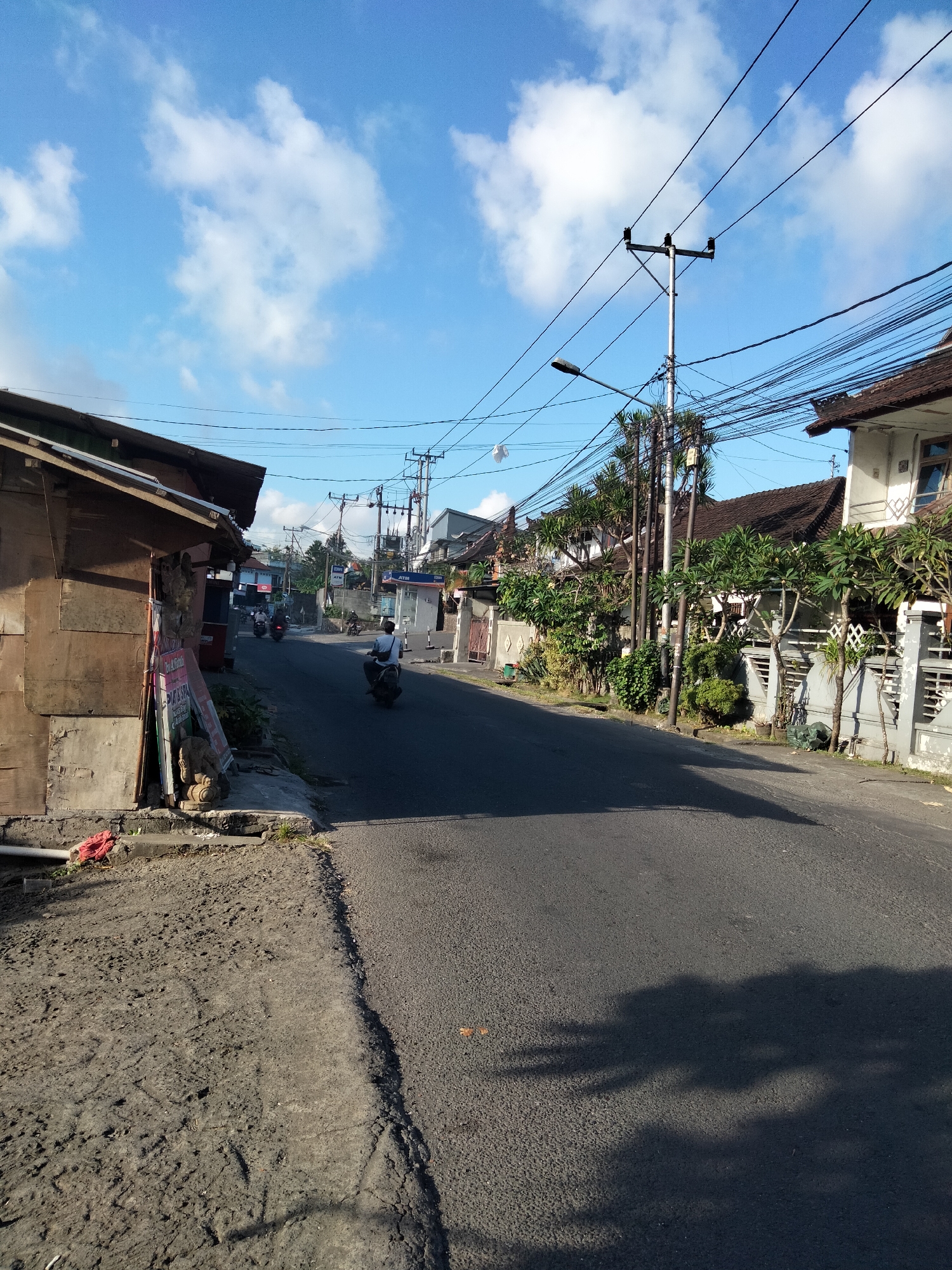 The image captures a sunlit street scene in Denpasar, Indonesia, likely in a residential or commercial area. The sky is a bright blue, dotted with fluffy white clouds. A paved road runs through the center of the frame, with a person riding a motorcycle in the distance, heading away from the viewer. To the left of the road is a weathered wooden structure, possibly a shop or residence, with posters and debris on the ground in front of it. To the right of the road, there are more modern buildings with tiled roofs, lush green trees, and a concrete wall with decorative fencing. Numerous electrical wires are strung overhead, supported by utility poles, indicating the presence of infrastructure. The overall atmosphere is calm and ordinary, depicting a typical day on a street in Denpasar.