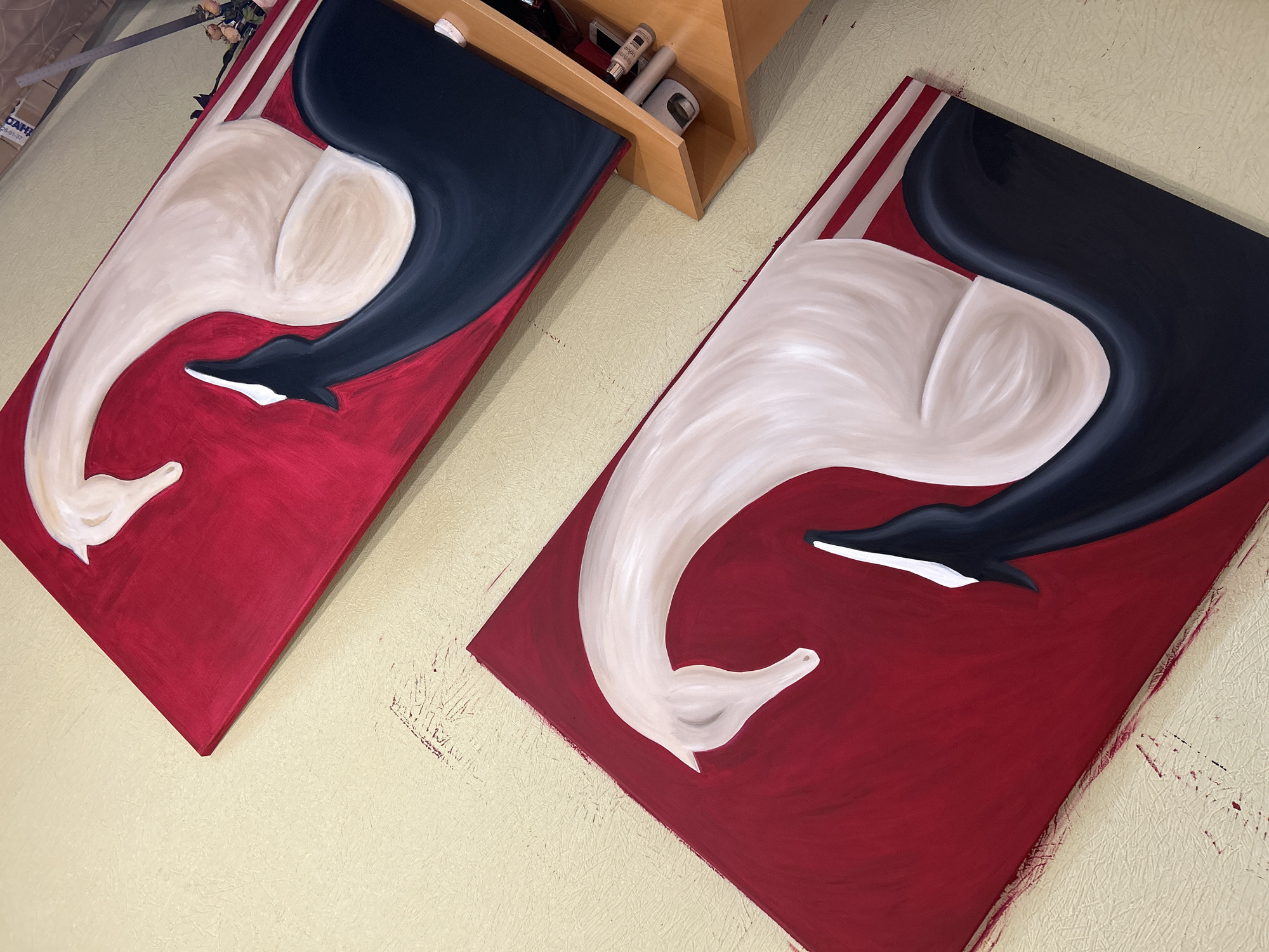 The image displays two abstract paintings, each featuring a stylized swan-like figure in white and black against a vibrant red background. The paintings are hung on a light-colored textured wall. The top painting is positioned diagonally from the top right to the bottom left, with its top edge partially obscured by a wooden shelf unit. The bottom painting is positioned similarly, below and to the left of the top one. The shelf unit, located to the right of the paintings, holds several items, including cylindrical objects and a phone. The scene appears to be an indoor setting, possibly an art studio or a room in a private residence, as indicated by the paintings and the shelf. There are no people visible in the image. The location is not specified in Moscow, Russia.