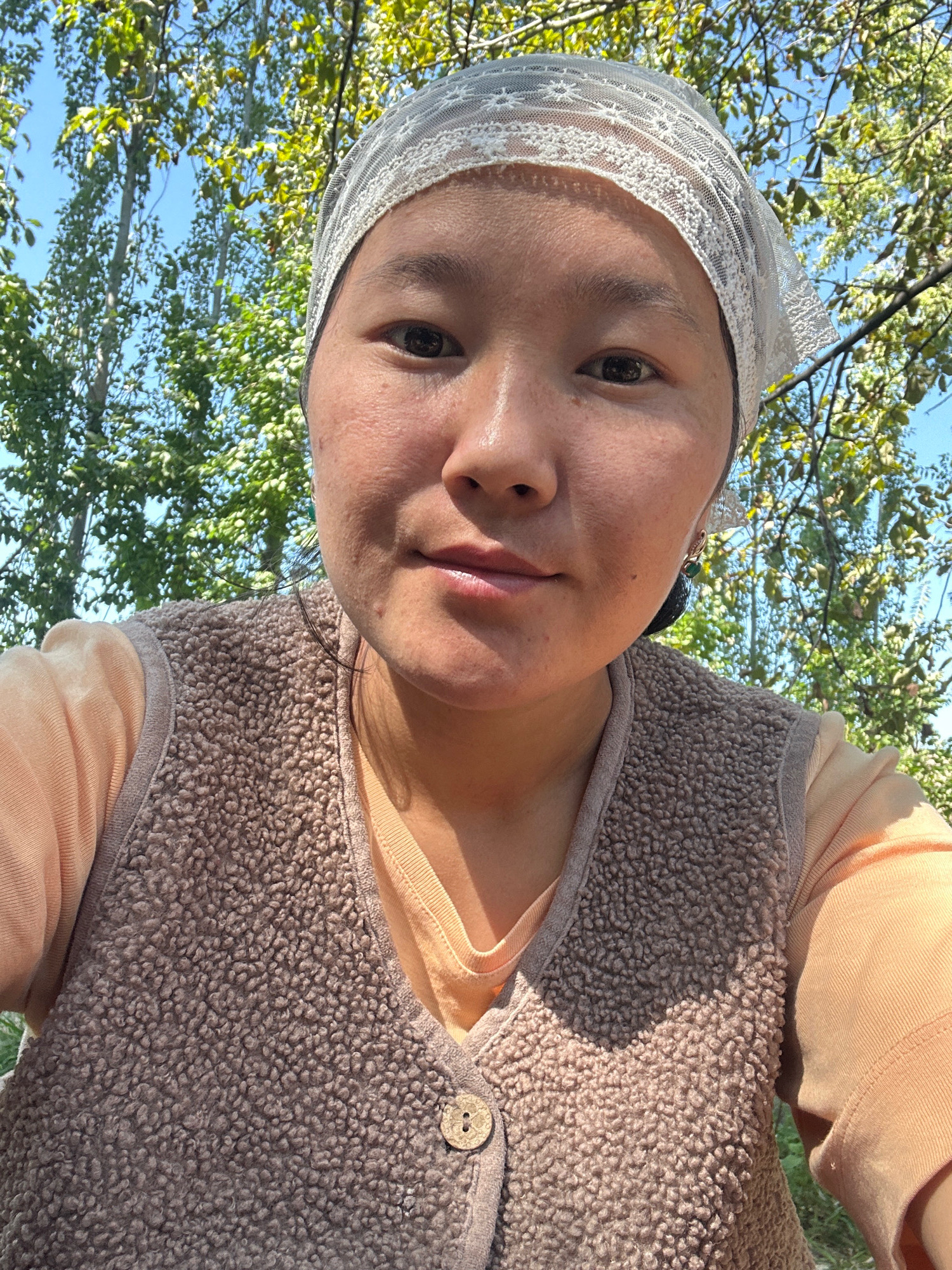 A close-up, front-facing selfie of a young woman wearing a light peach-colored long-sleeved shirt and a brown, fuzzy vest with a wooden button. She also wears a white, lacy headscarf. The woman has dark hair and dark eyes, and her skin appears clear with some freckles. She is outdoors with a backdrop of tall green trees and a clear blue sky, suggesting a location in Toktogul, Kyrgyzstan, likely during a warm season. The lighting suggests it is daytime with bright sunlight.