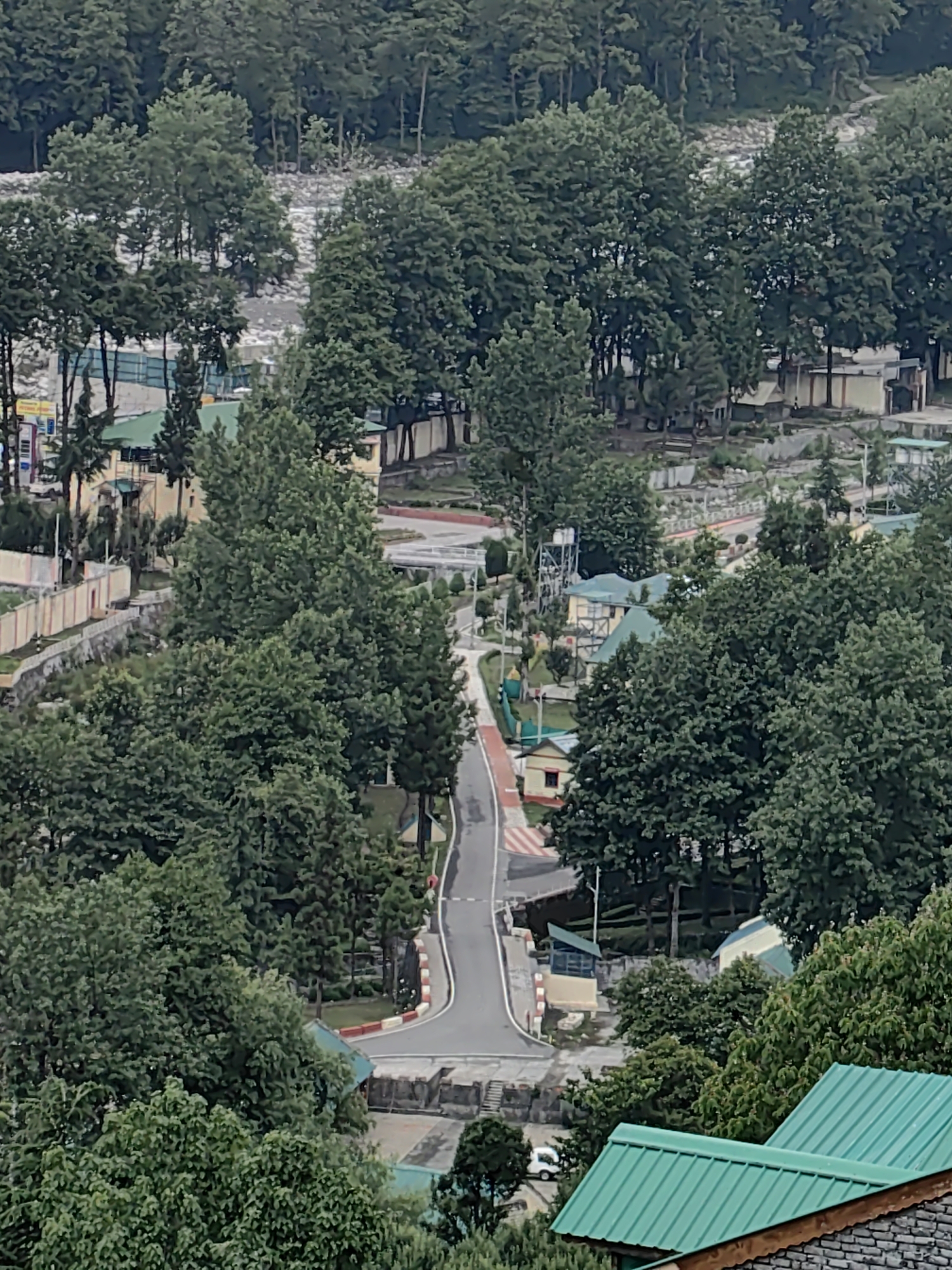 This is an aerial, high-angle shot of a mountainous landscape in Vashisht, India. A paved road winds through a dense forest of green trees, leading towards a cluster of buildings with green roofs. Some of the buildings appear to be houses or cabins, while others could be part of a larger complex or resort. A white car is visible on the road. In the background, a river or body of water is visible flowing through a rocky terrain, surrounded by more trees. The overall scene suggests a rural or semi-rural setting with abundant natural beauty.