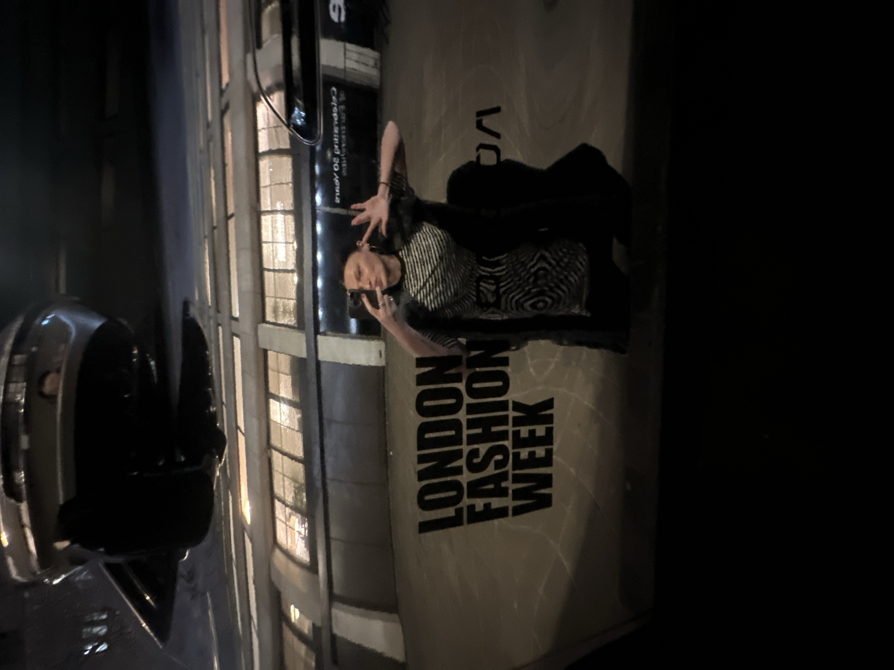 A person poses in front of a "London Fashion Week" sign at night. The individual has short hair, wears a striped shirt, and is seen with raised hands. A modern car is parked on the street. A building with multiple windows is visible in the background. The scene is dimly lit, with the sign and building illuminated against the dark surroundings.