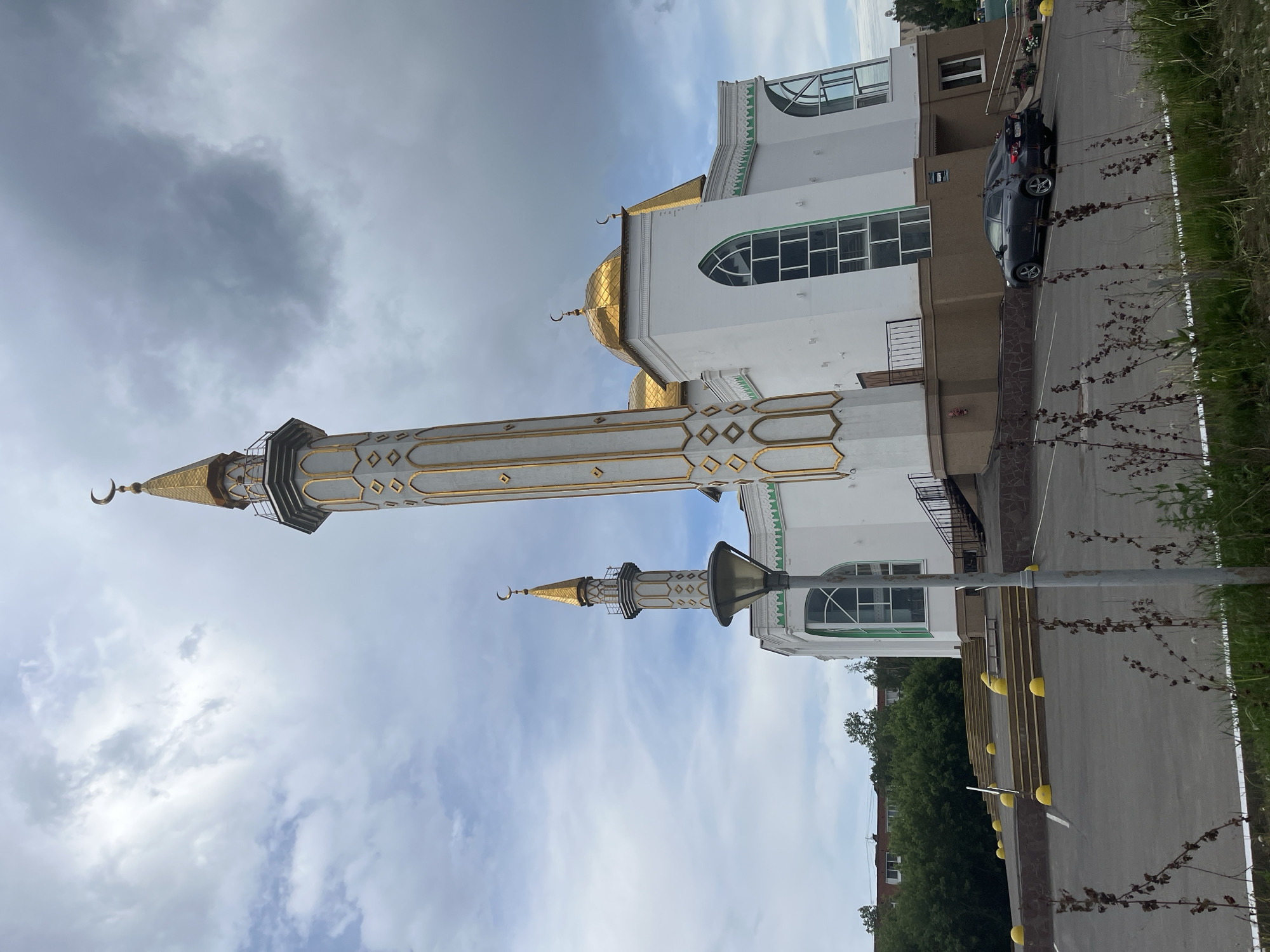 The image shows a mosque in Izhevsk, Russia. The mosque is white with gold domes and minarets. A dark-colored car is parked nearby. The sky is cloudy. There are trees and other vegetation in the background. A street is also visible with markers.